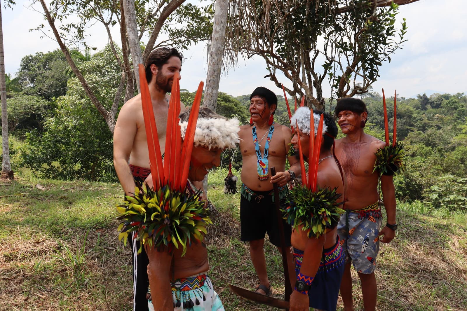 Dressed by the community, laughing with the community. Some invitations you just say yes to. A smiling Western traveler wearing a traditional feathered headdress with tall red feathers and leaf adornments stands among Yanomami men during a Pico da Neblina Expedition cultural encounter, surrounded by Amazon forest