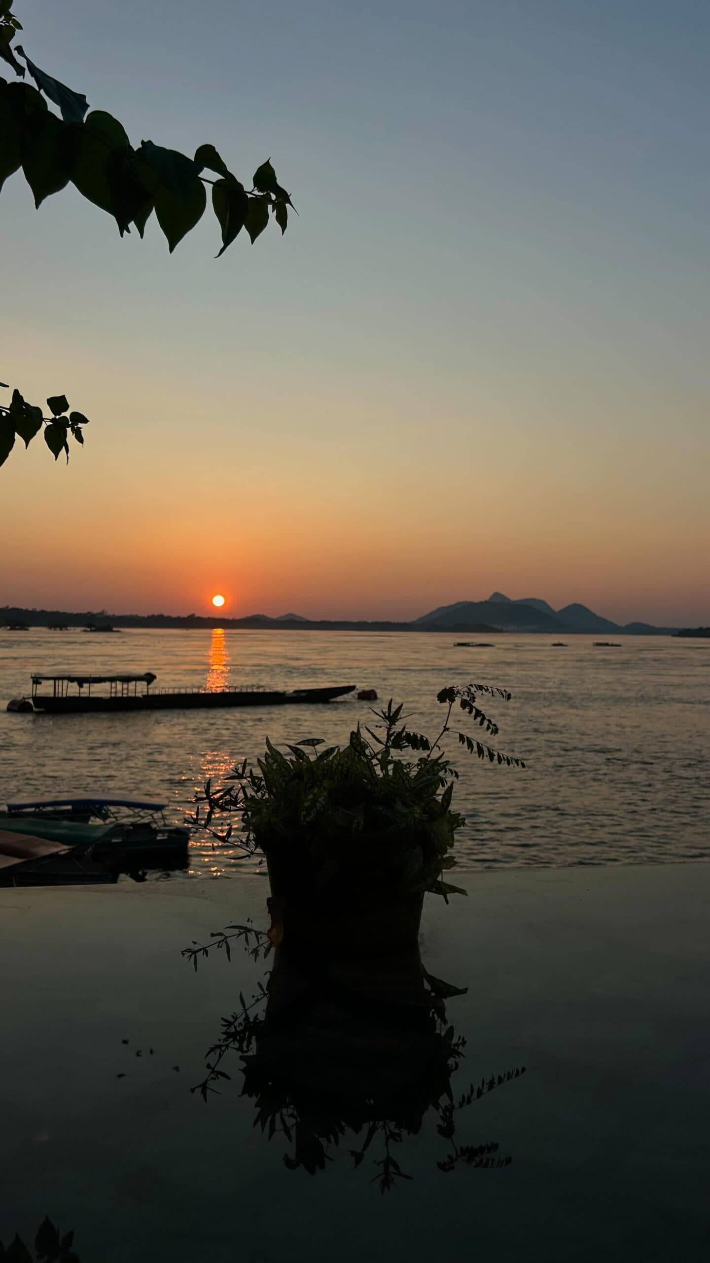 Some evenings the river earns its keep, just sitting there looking like that. A glowing orange sun sets over a wide river on the Pico da Neblina Expedition, with silhouetted boats on the water, a potted plant in the foreground, and distant hills on the horizon