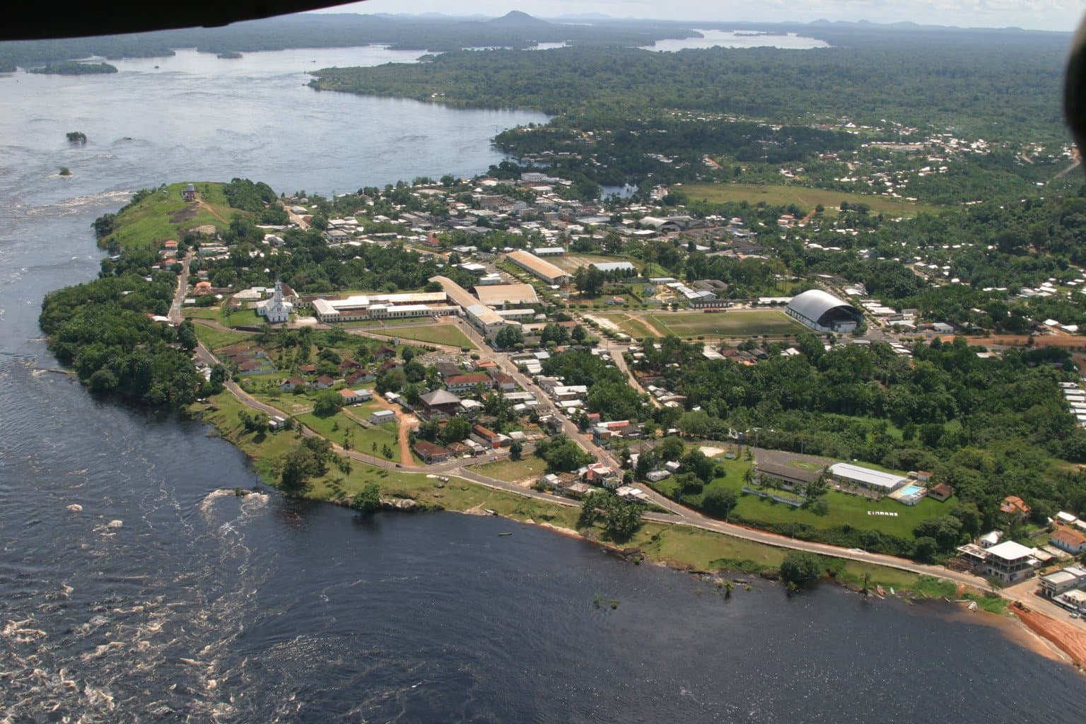São Gabriel da Cachoeira from the air: a small town at the edge of everything. Aerial view of São Gabriel da Cachoeira, the starting point of the Pico da Neblina Expedition, showing the town's white church, grid of streets and buildings on a forested peninsula surrounded by the dark waters of the Rio Negro, with rainforest and rocky outcrops stretching to the horizon