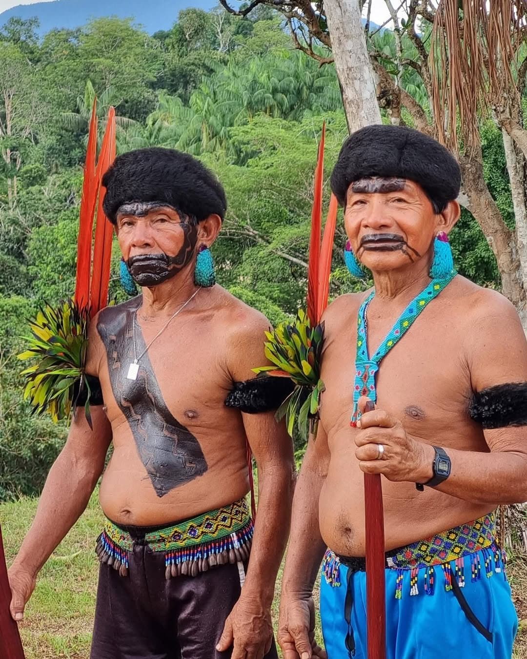 Before the first step, the forest gets its say. Two Yanomami elder men in traditional ceremonial dress — black body paint, red feather headdresses, turquoise bead earrings and woven bead belts — standing together before the Pico da Neblina Expedition trek begins, with lush rainforest in the background