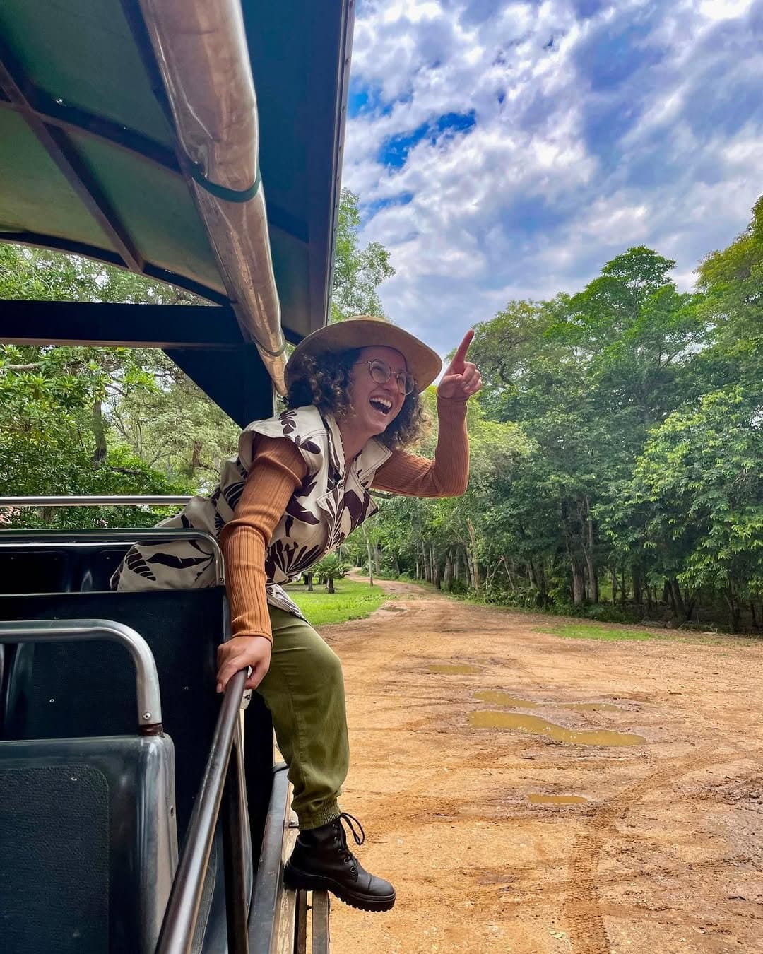 Spotted something, the finger goes up and the whole truck stops. A laughing woman in a sun hat leans out of an open 4x4 vehicle on a southern pantanal safari, pointing skyward along a muddy dirt road lined with dense green trees.