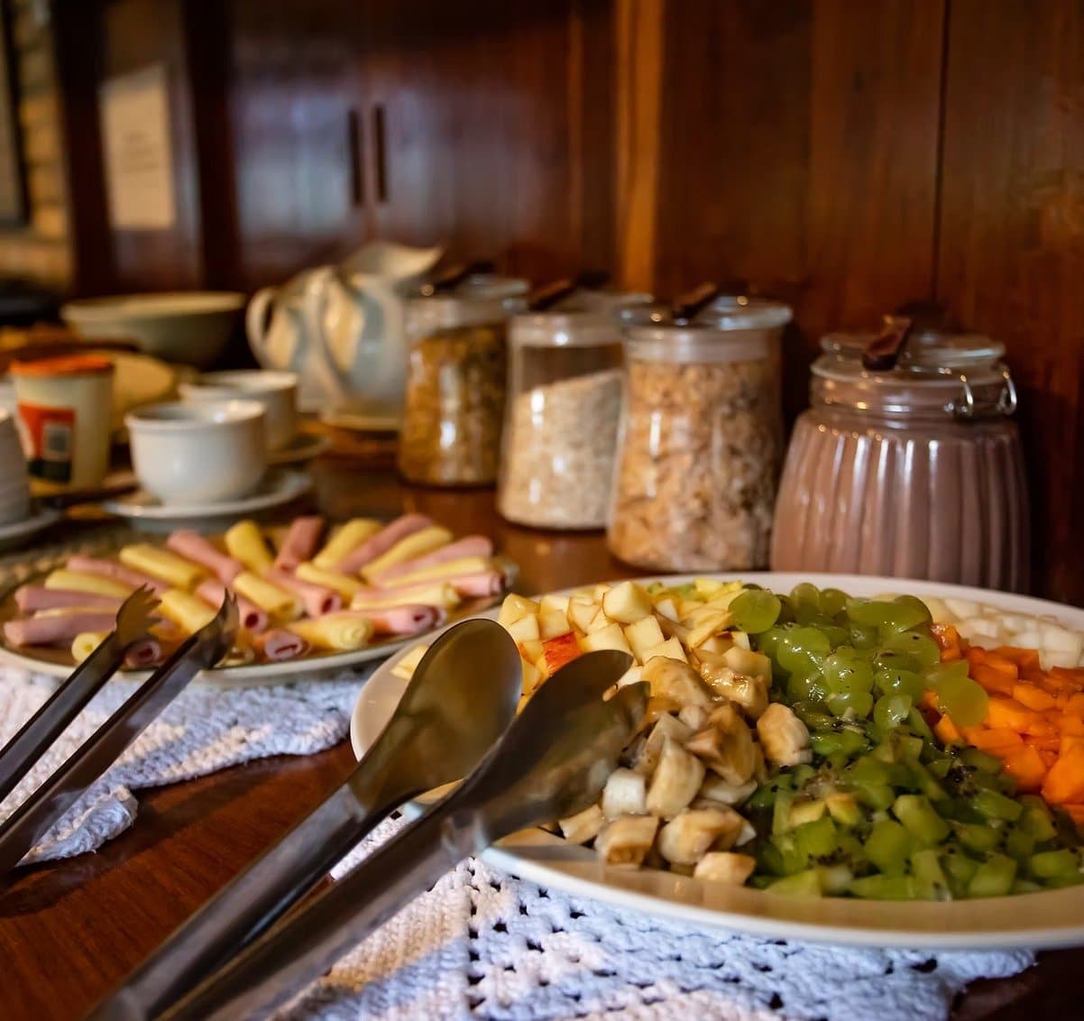 Fuel up well before the anteaters get a head start on you. A southern pantanal safari lodge breakfast buffet spread with sliced fresh fruit including banana, kiwi, grapes and papaya, rolled cold cuts and cheese, granola jars, ceramic cups and a pitcher on a dark wood sideboard