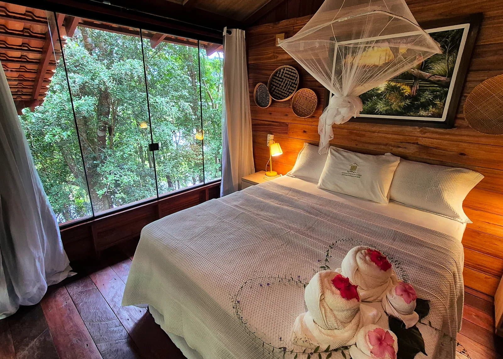 Towels shaped like swans, forest an arm's reach away. Sleep well. Cozy Amazon boutique lodge bedroom with a mosquito-net canopy bed, warm timber walls, woven basket decor, towels folded into flowers, and a floor-to-ceiling window overlooking dense green forest canopy