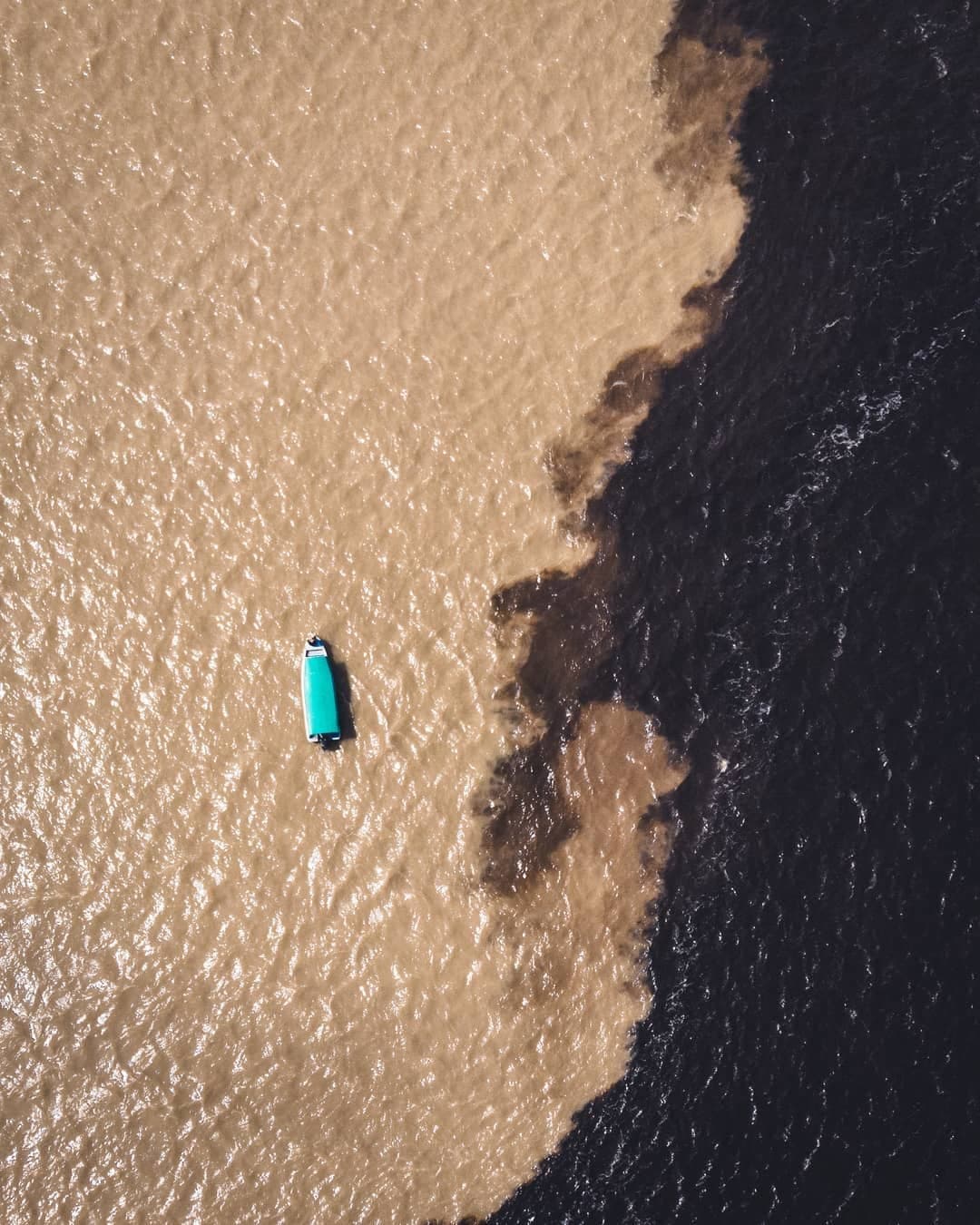 One boat, two rivers, zero hesitation. Aerial view of a turquoise-roofed boat crossing the Amazon jungle cruise Brazil highlight — the Meeting of the Waters, where sediment-rich tan water meets dark black water in a dramatic natural boundary