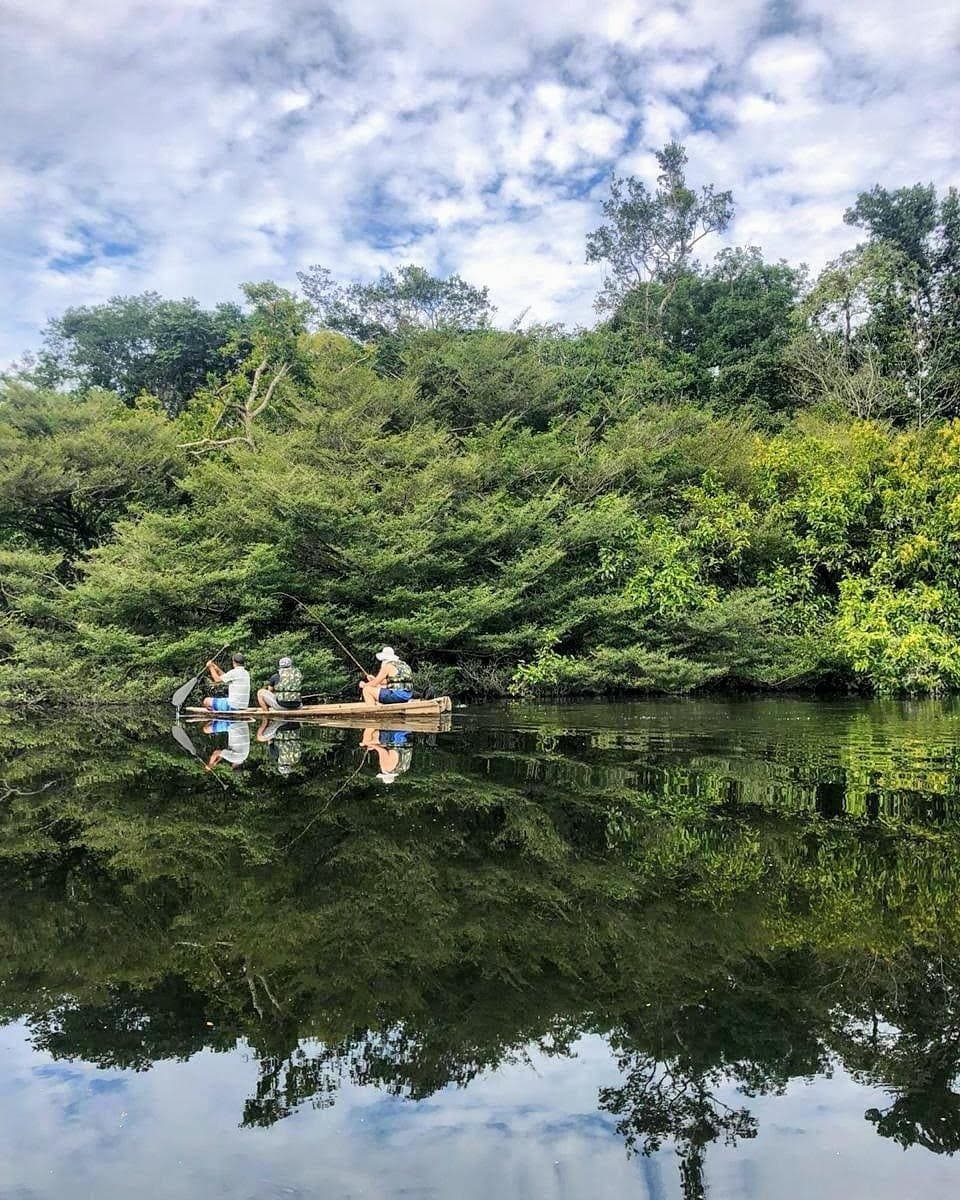 Lines in the water, forest all around, and a piranha somewhere below with opinions. Three guests fishing from a wooden canoe on a calm, mirror-still river during an amazon river cruise brazil, surrounded by dense tropical forest under a partly cloudy sky
