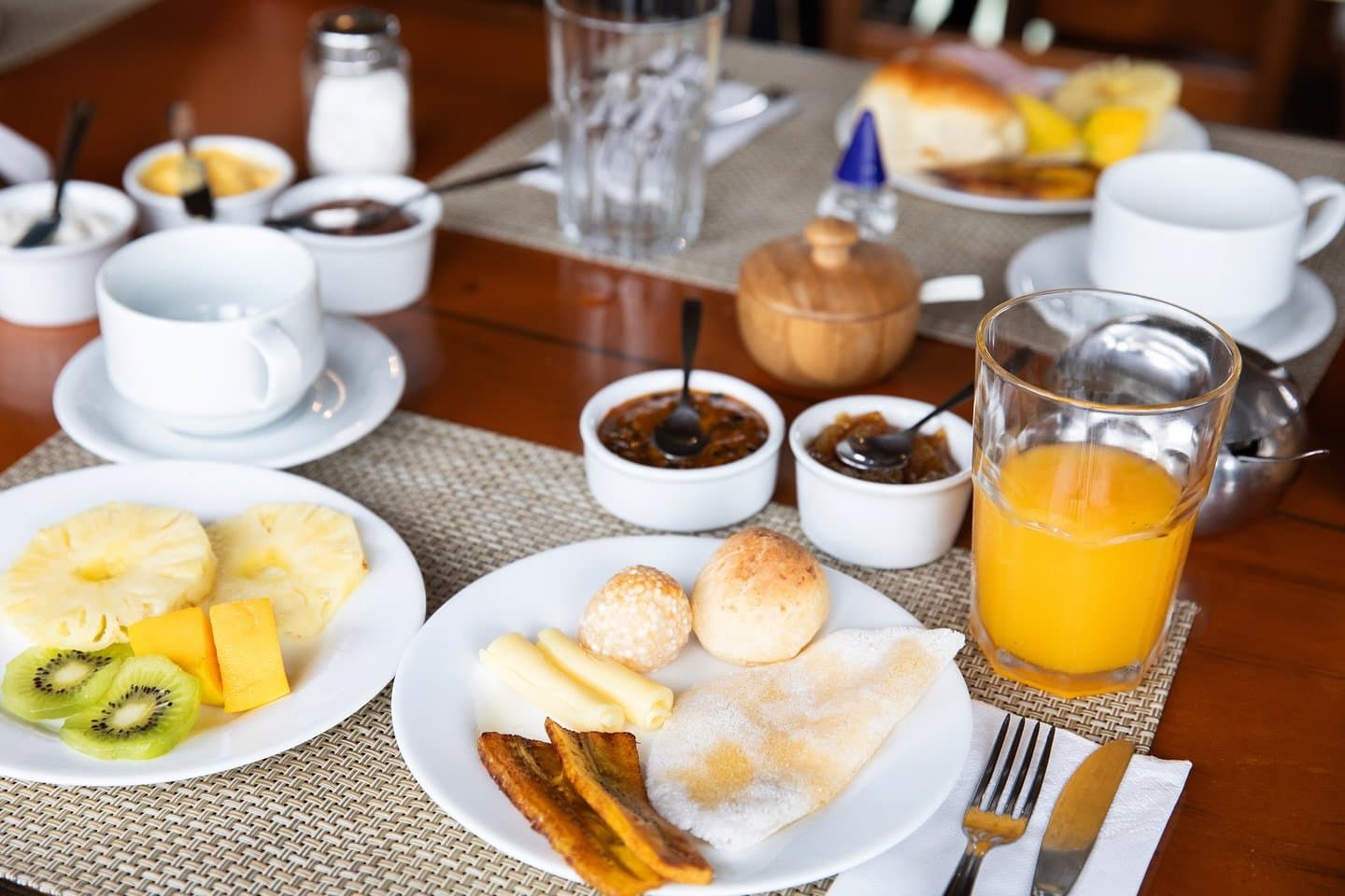 Fuel up properly before a morning on the river. A full amazon river cruise brazil breakfast spread with pão de queijo, tapioca, fried plantain, sliced cheese, fresh pineapple, kiwi, orange juice, fruit preserves, and coffee cups set on a woven placemat