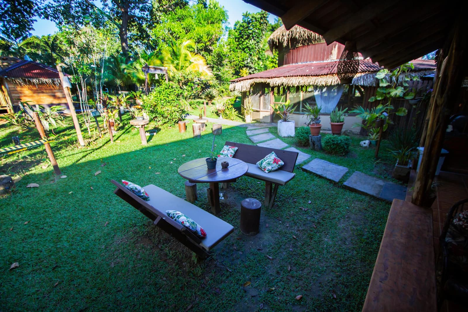 The kind of garden that makes you forget you had plans. Cushioned wooden benches and a round table arranged on a green lawn at the Amazon Turtle Lodge, with thatched-roof bungalows and lush tropical garden in the background