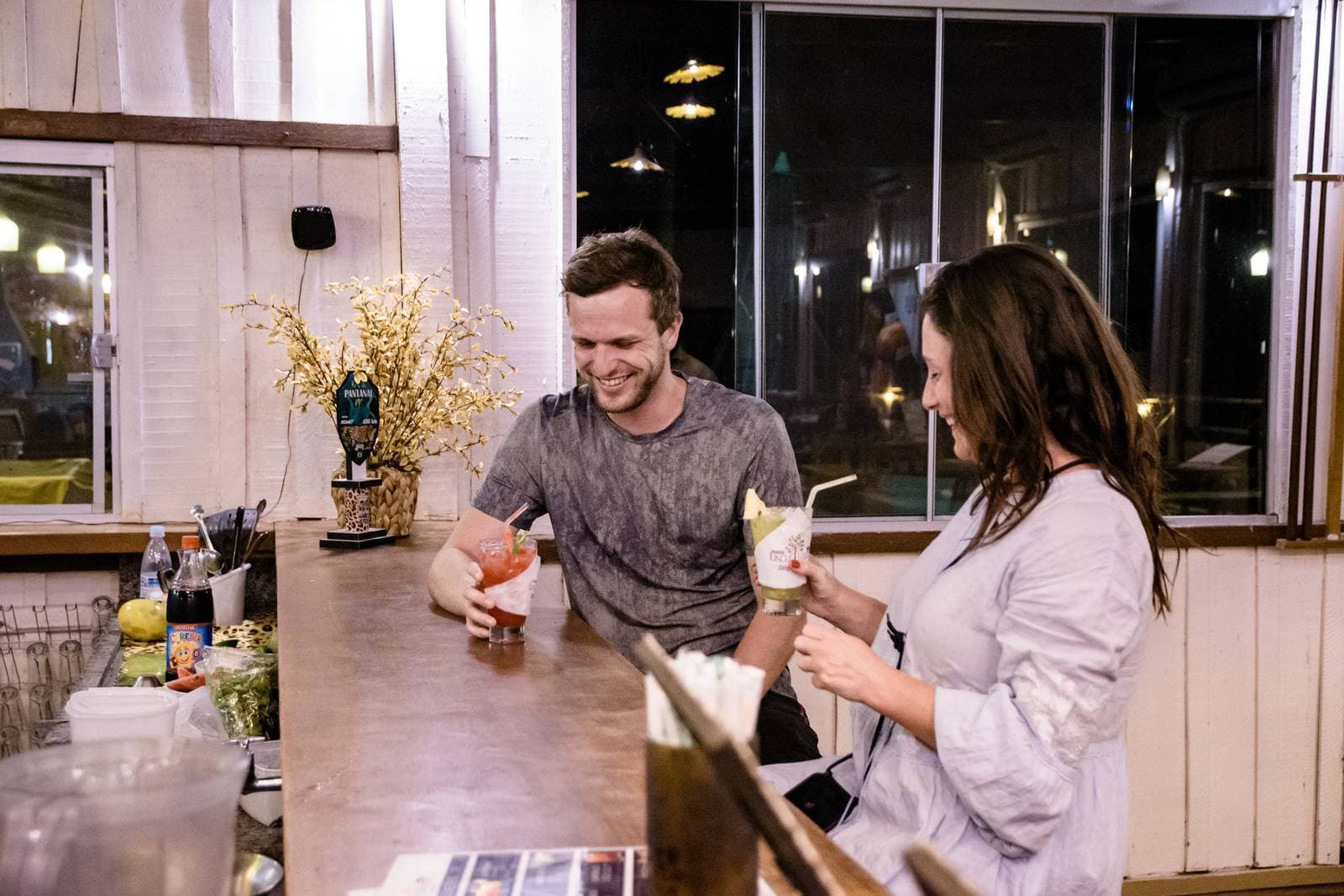 The best Pantanal stories start right here, drinks in hand. Two travelers laughing and holding cocktails at a rustic indoor bar counter on a budget Pantanal tour, with warm lighting and a Pantanal beer tap visible in the background