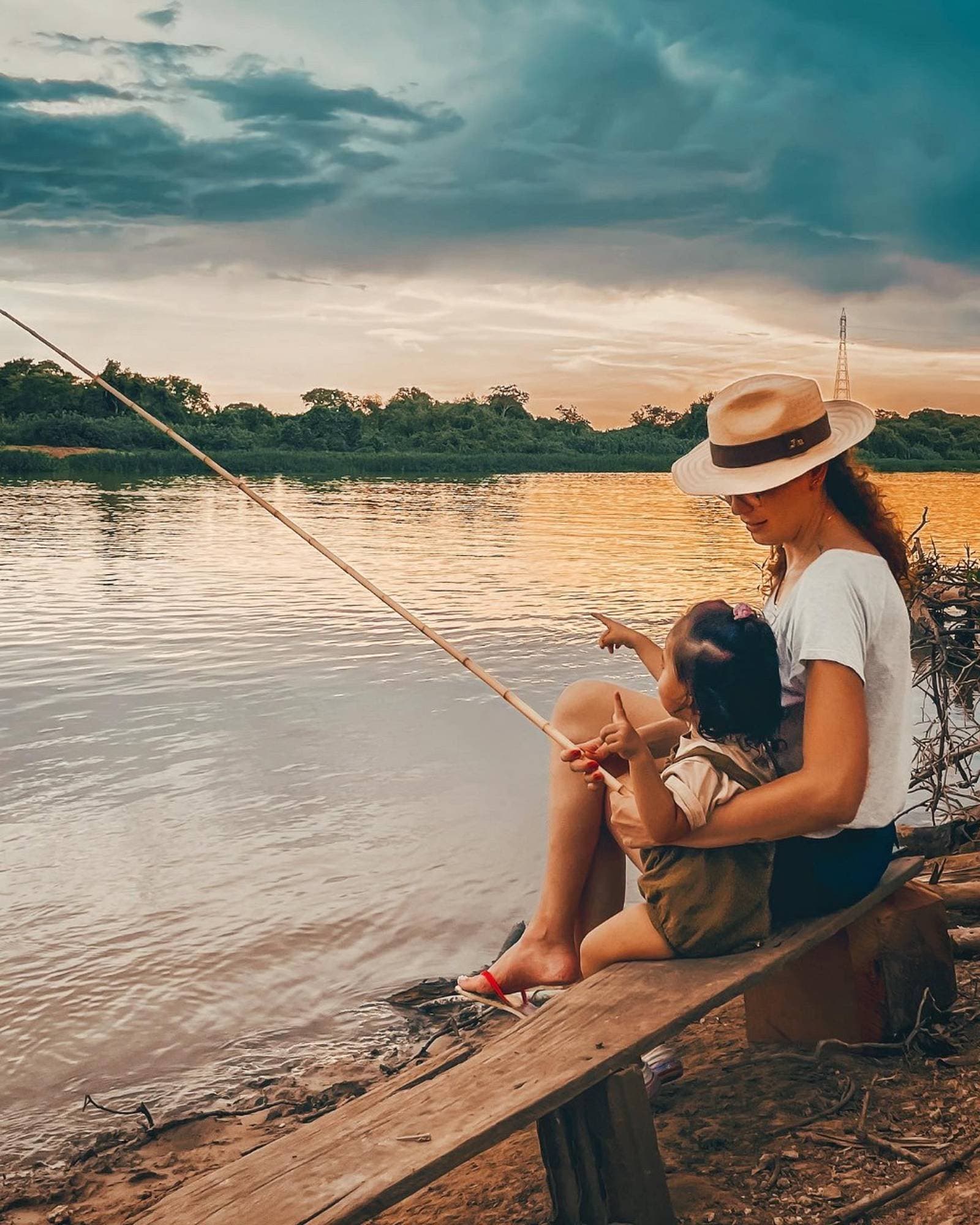The river teaches patience. So does a toddler with a fishing rod. A woman in a panama hat holds a toddler on her lap while fishing with a bamboo rod on the Miranda River at sunset during a budget Pantanal tour, golden light reflecting off the calm water