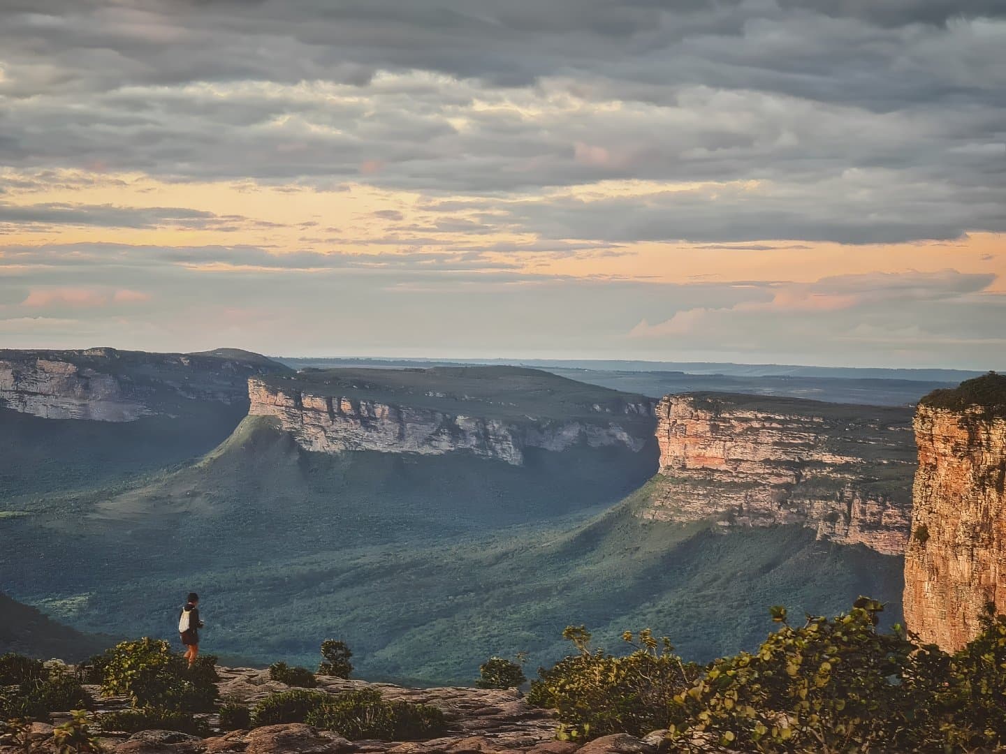 That moment when the canyon does all the talking and you just listen. A lone hiker stands on a rocky ledge overlooking the vast sandstone tablelands and green valleys of a Chapada Diamantina trip at dusk, with warm light breaking through dramatic clouds