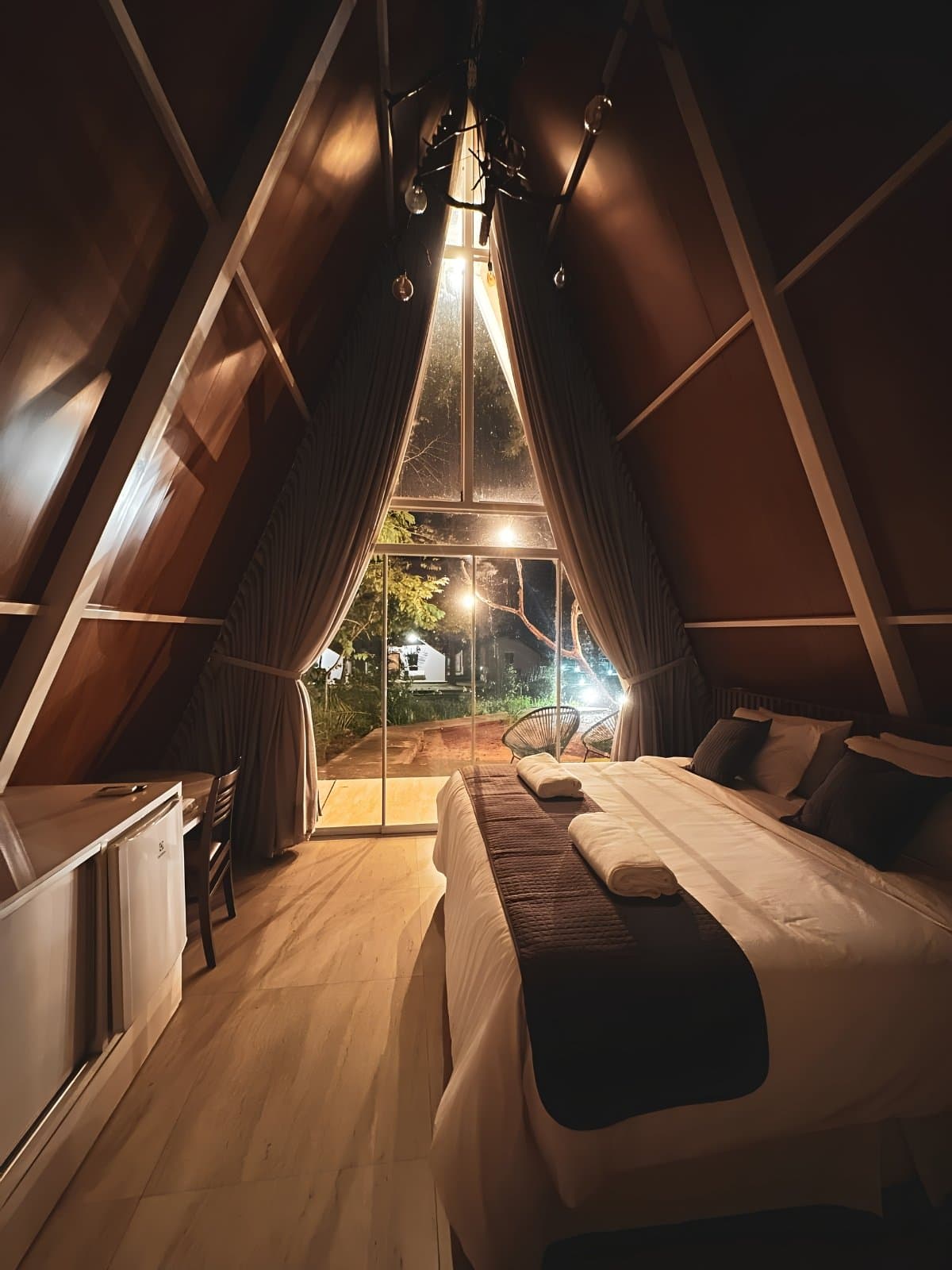 Warm wood, soft light, and the Cerrado just outside the glass. Interior of an A-frame wooden cabin at night on a Jalapão adventure, featuring a double bed with folded towels, warm string lights, and a floor-to-ceiling triangular window overlooking a lit garden