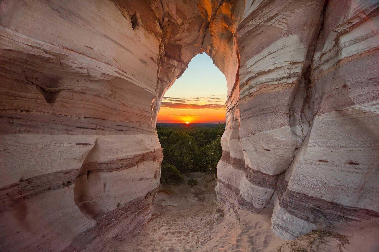 Ancient rock, one perfect hole, and the sun decides to put on a show. View through a natural sandstone arch at Pedra Furada, framing a fiery orange sunset over a green Cerrado canopy during a Jalapão adventure