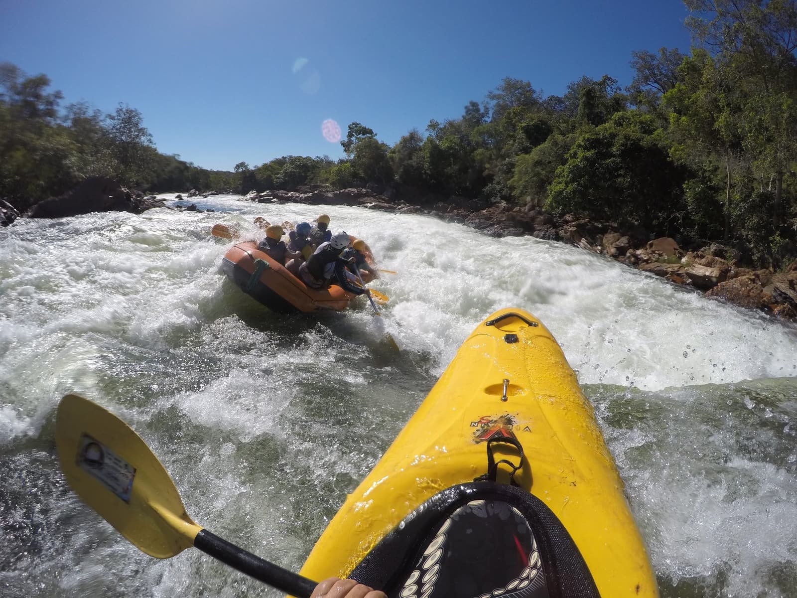 Front-row seat on the Novo River, and the raft crew is having none of your calm. Point-of-view shot from a yellow kayak during Jalapão trekking, with a crew in an orange raft powering through churning Class IV whitewater rapids, green-forested riverbanks under a clear blue sky