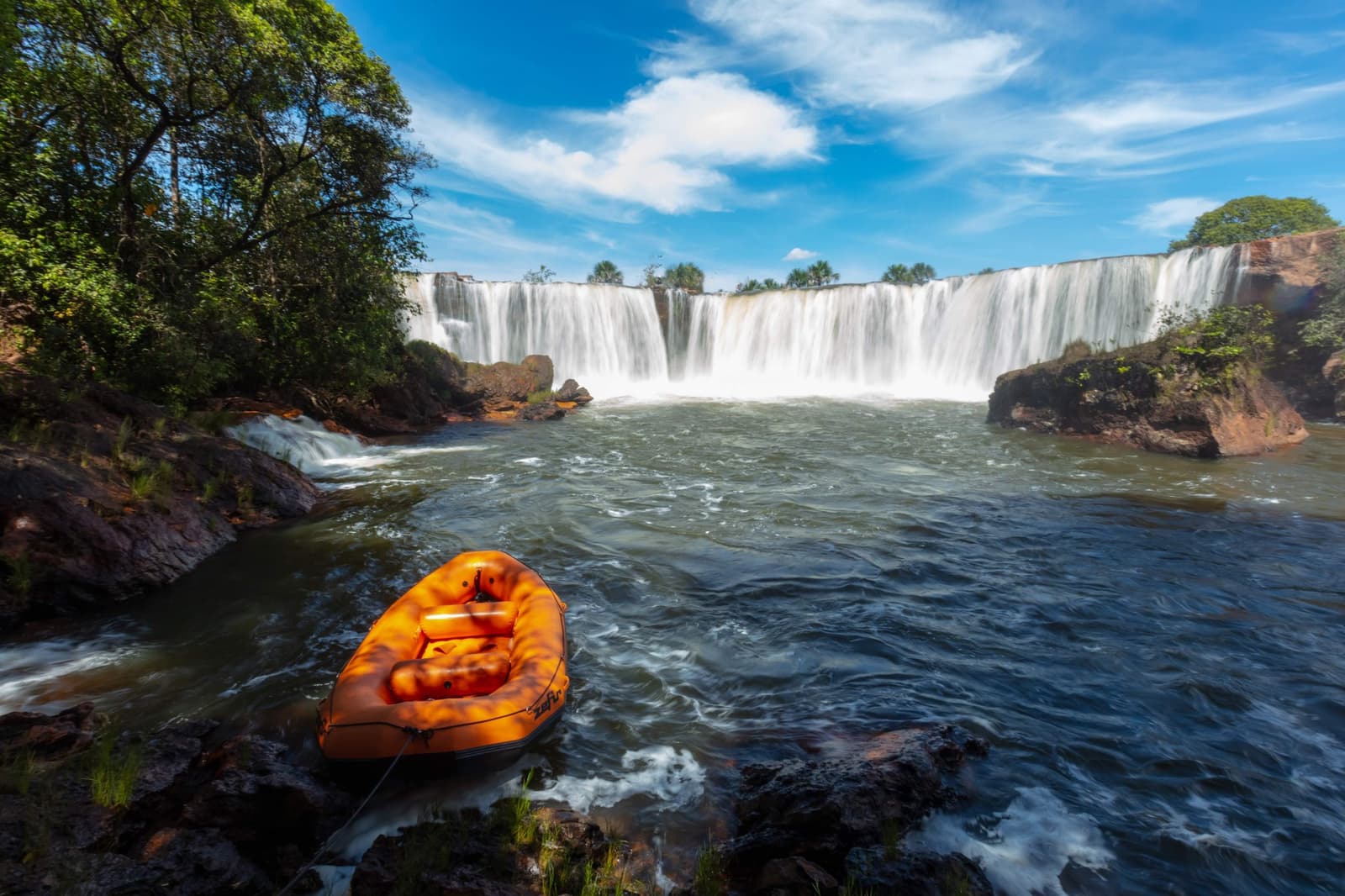 The raft waits, the waterfall does not. Pick your moment. Photo: Alberto Andrich An orange inflatable raft moored on rocky shore in front of a wide curtain waterfall during a Jalapão trip, with green cerrado forest framing a blue sky above