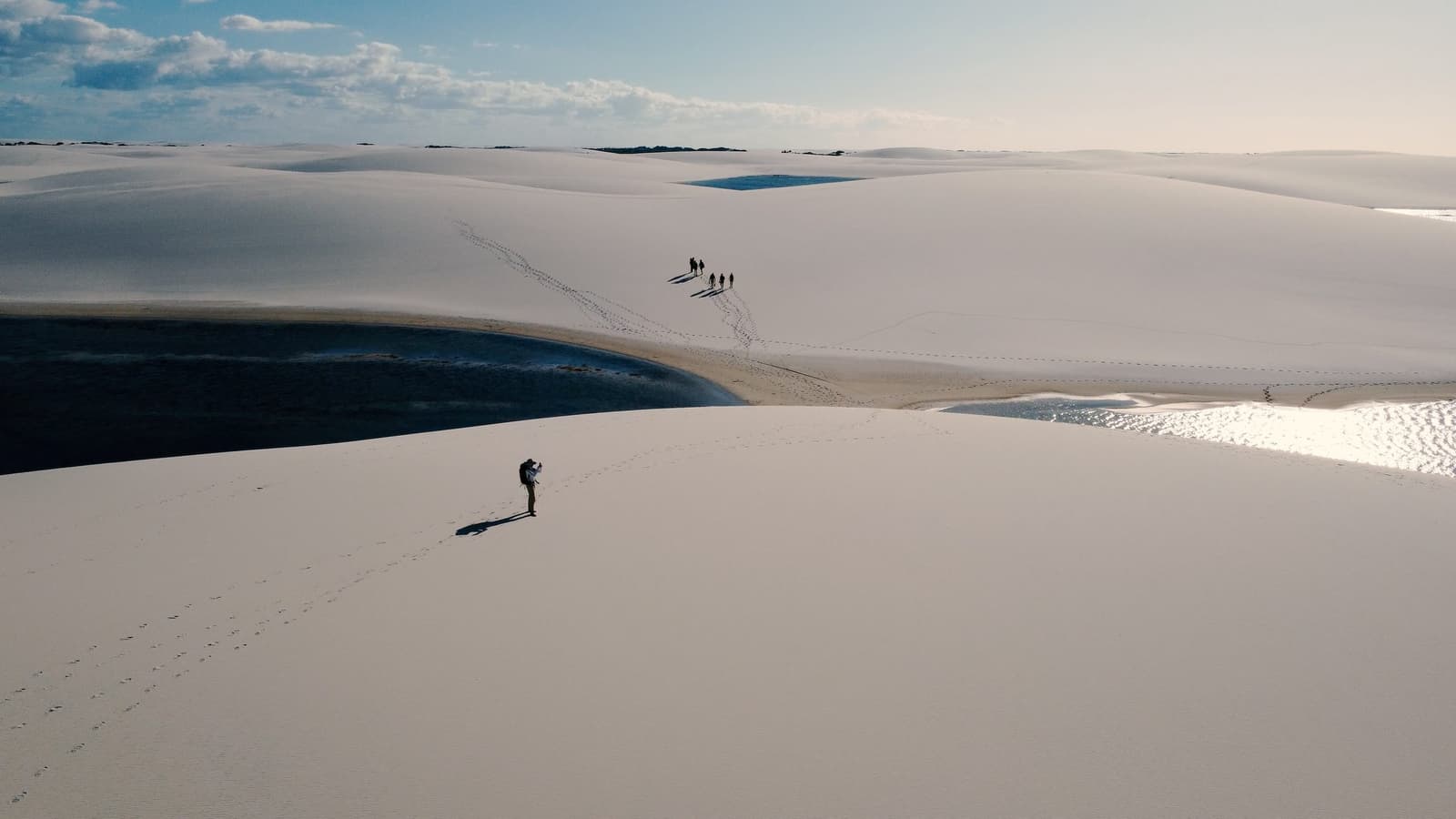 Out here, the dunes swallow distance whole, and you realise how small a good adventure makes you feel. Aerial view of trekkers leaving footprints across vast white dunes in the Lençóis Maranhenses Full Itinerary, with dark blue lagoons nestled between the sandy ridges under a partly cloudy sky