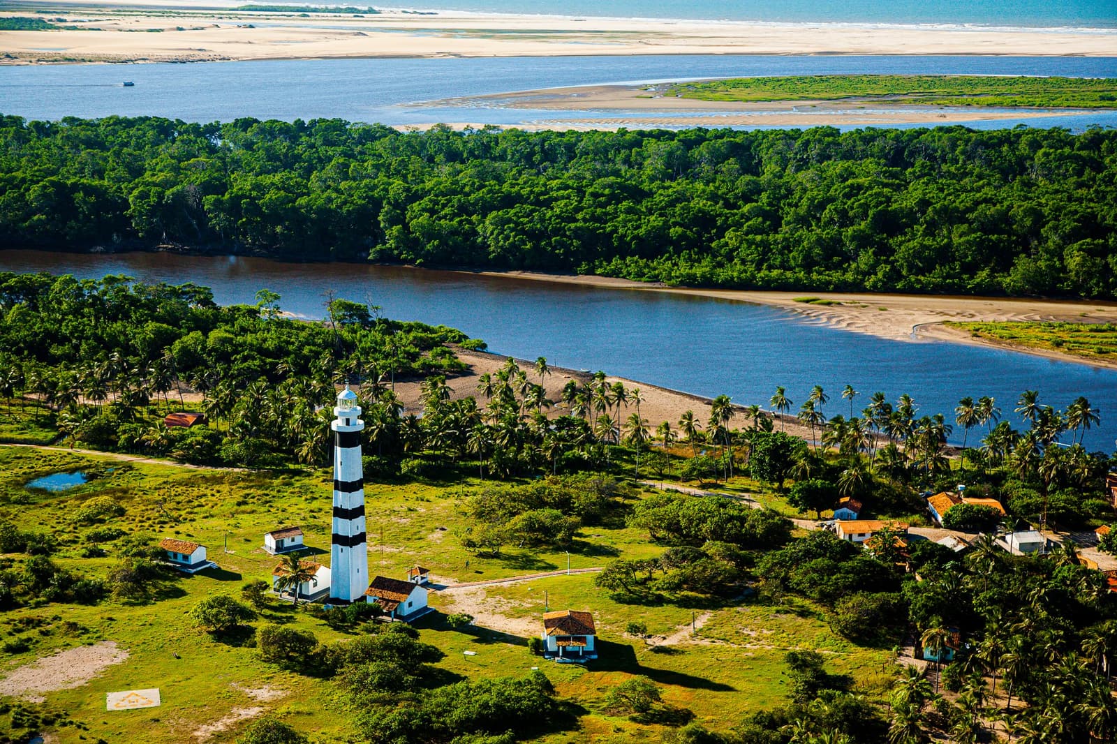 The lighthouse has been keeping watch here long before tourism found this corner of Maranhão. Aerial view of a black-and-white striped lighthouse surrounded by palm trees and small buildings, with the Preguiças River, dense forest, and white sand dunes stretching to the horizon — a landmark stop on the Lençóis Maranhenses Full Itinerary