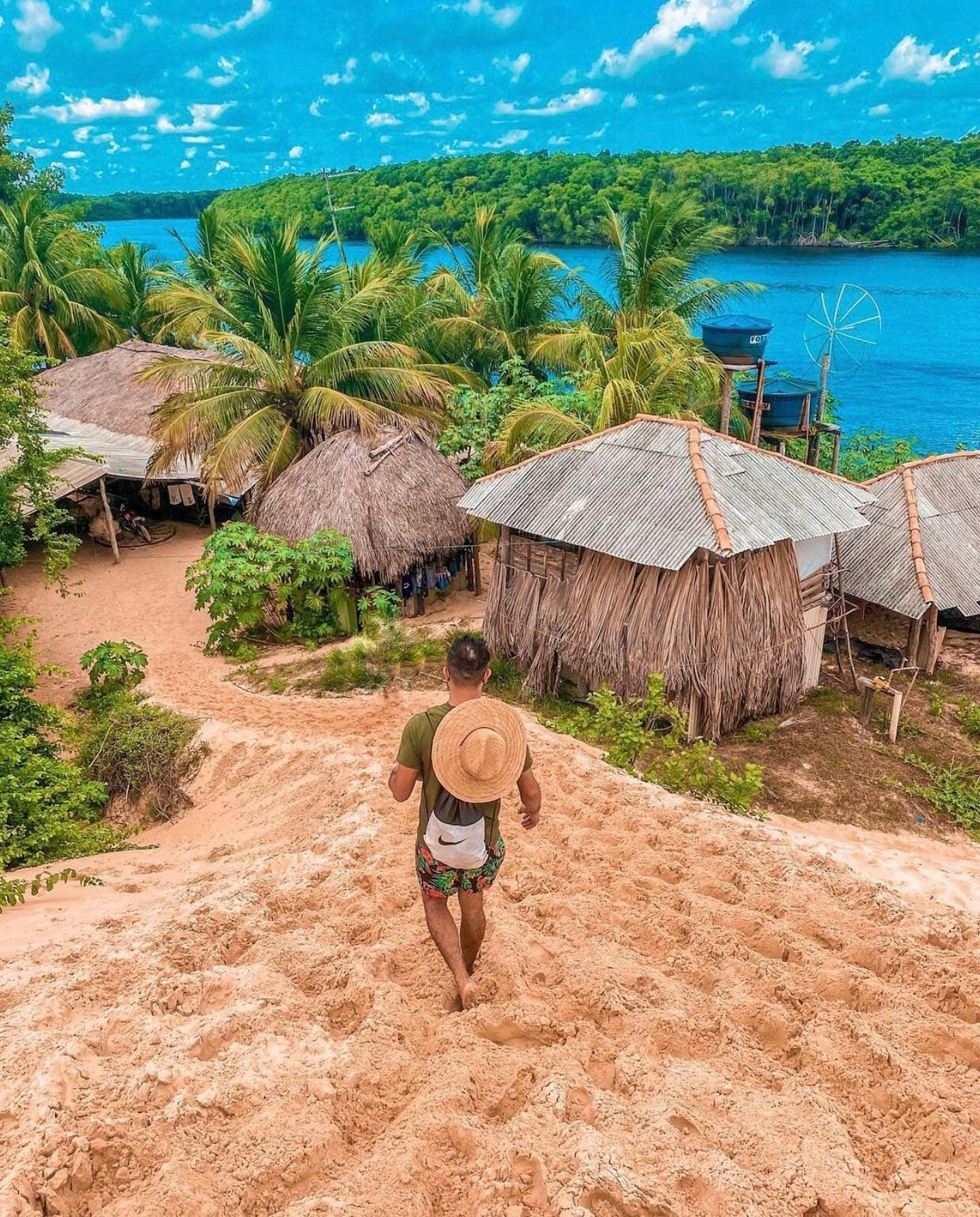 Some paths don't need a sign, just follow the sand down. A traveller with a straw hat and small backpack walks barefoot down a sandy slope toward a cluster of thatched and tin-roofed huts surrounded by palm trees and a wide blue river on the Lençóis Maranhenses hike