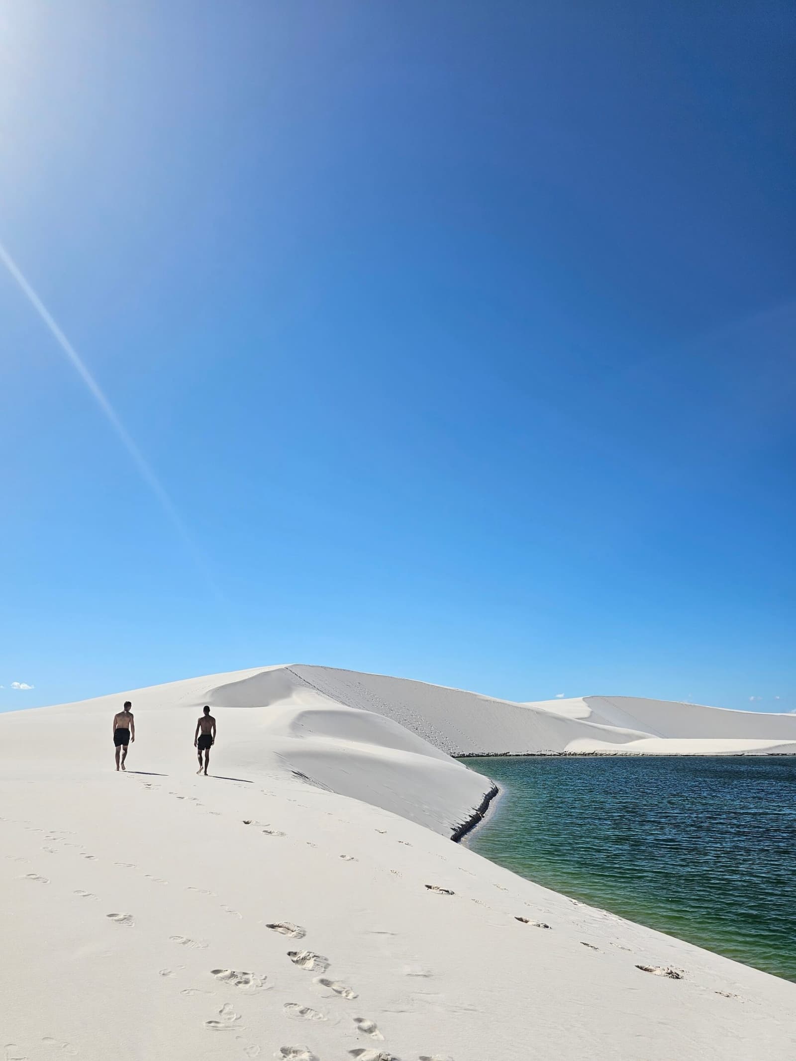 Just two people, an absurd amount of white sand, and water that has no business being that green. Two hikers walking barefoot up a vast white sand dune beside a clear green lagoon during a Lençóis Maranhenses hike, under a deep blue midday sky