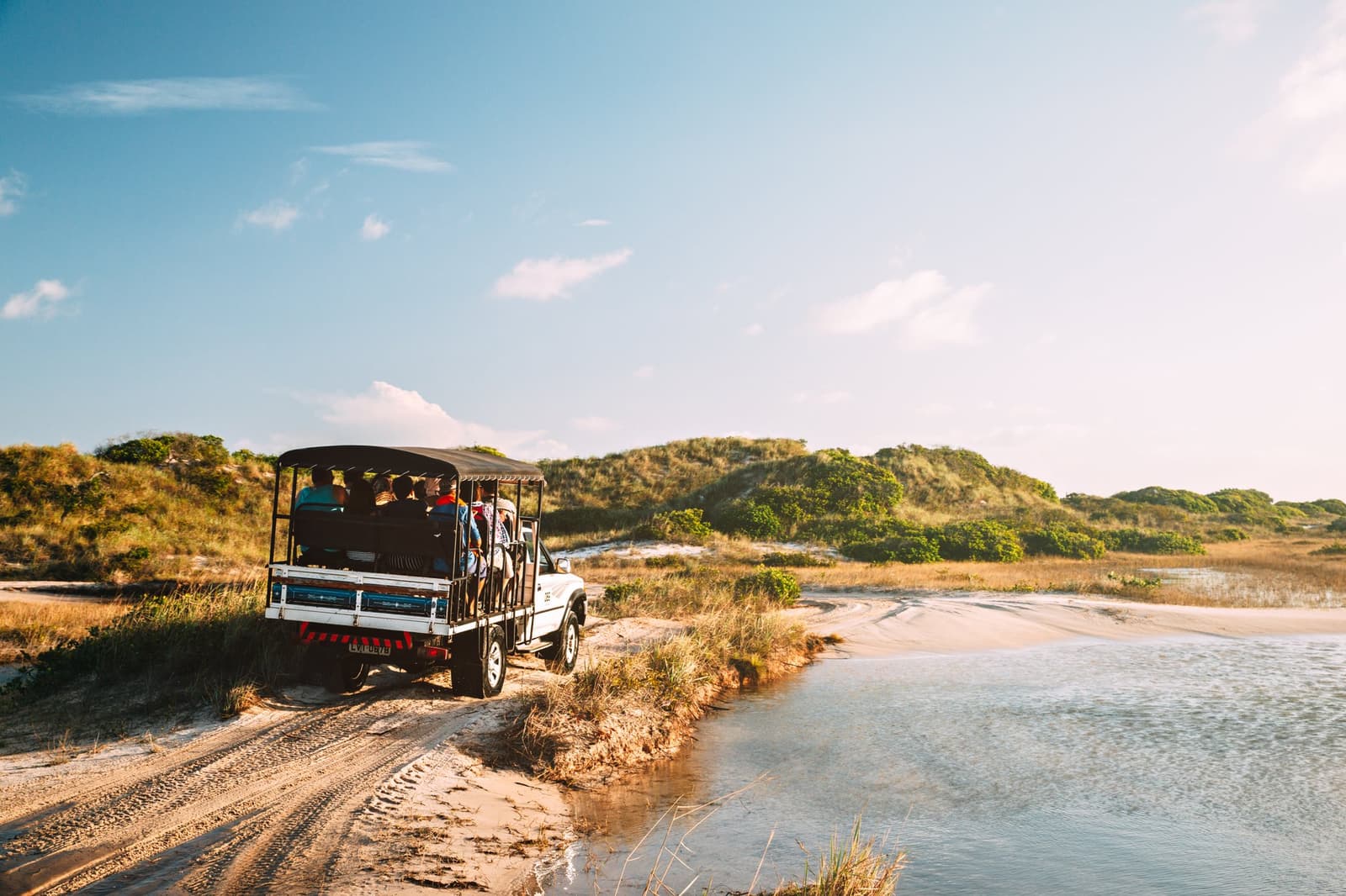 The only traffic jam out here is deciding which lagoon to stop at first. An open-top 4x4 vehicle carrying a group of visitors along a sandy track beside a shallow lagoon, with vegetated dunes rising under a blue sky in Lençóis Maranhenses Santo Amaro