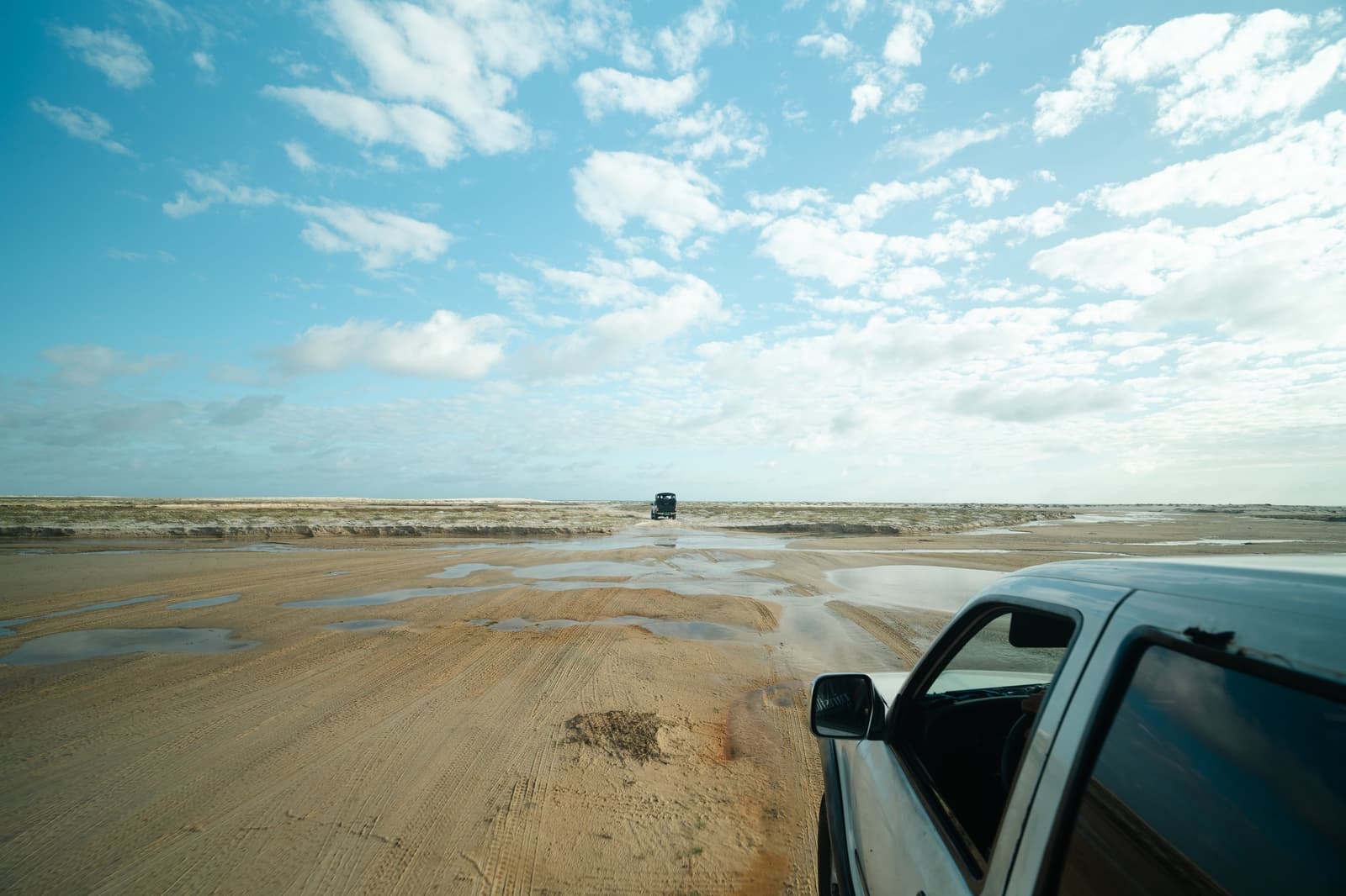 Out here, the road is whatever the tide left behind. Two 4x4 vehicles crossing a wide expanse of wet sandy flats under a partly cloudy blue sky during the Lençóis Maranhenses trek from Atins, with shallow pools of water reflecting the light across the terrain