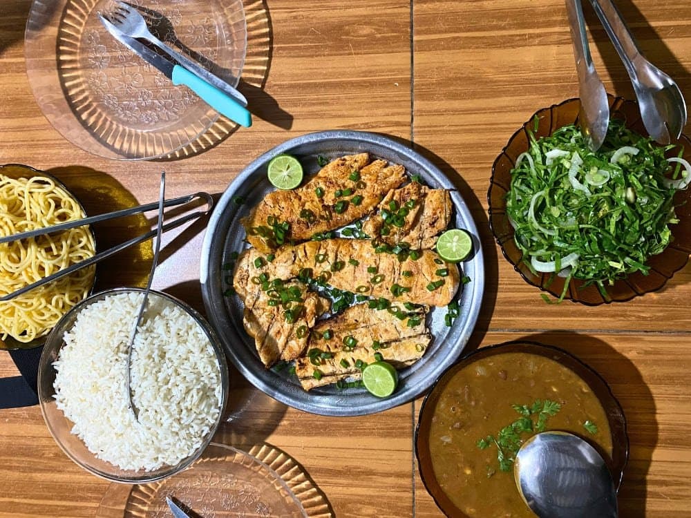 Lunch here is a full argument for eating with your hands. Overhead view of a rustic meal spread on a wooden table during a lencois maranhenses trek: a metal platter of grilled fish fillets topped with sliced green onion and lime, bowls of white rice, noodles, green salad, and a brown bean stew with cilantro