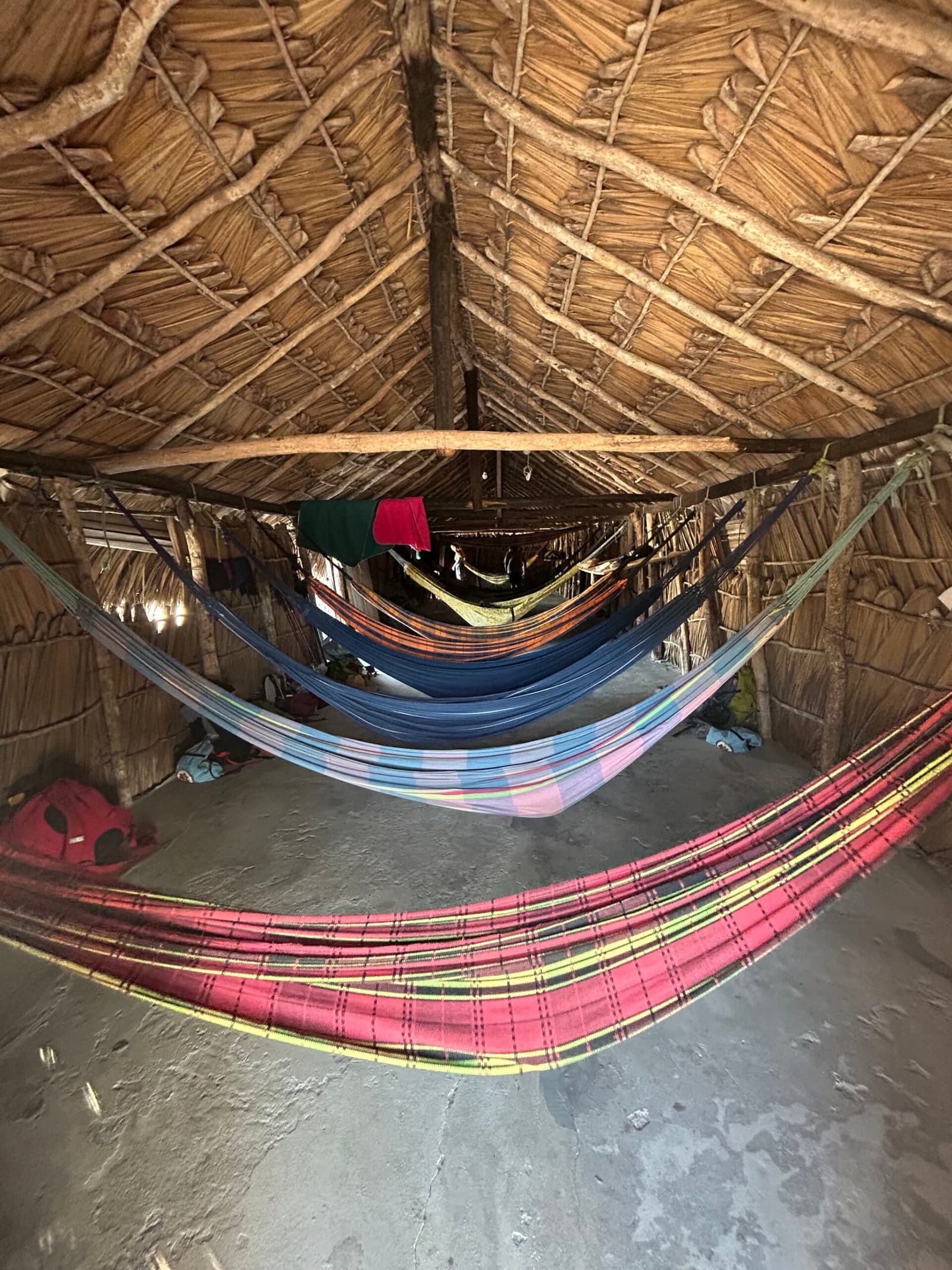 Your bed tonight: a hammock, a thatched roof, and no complaints. Multiple colorful hammocks strung inside a rustic thatched-roof shelter during the lencois maranhenses trek, with backpacks resting on the earthen floor beneath a lattice of raw wooden beams