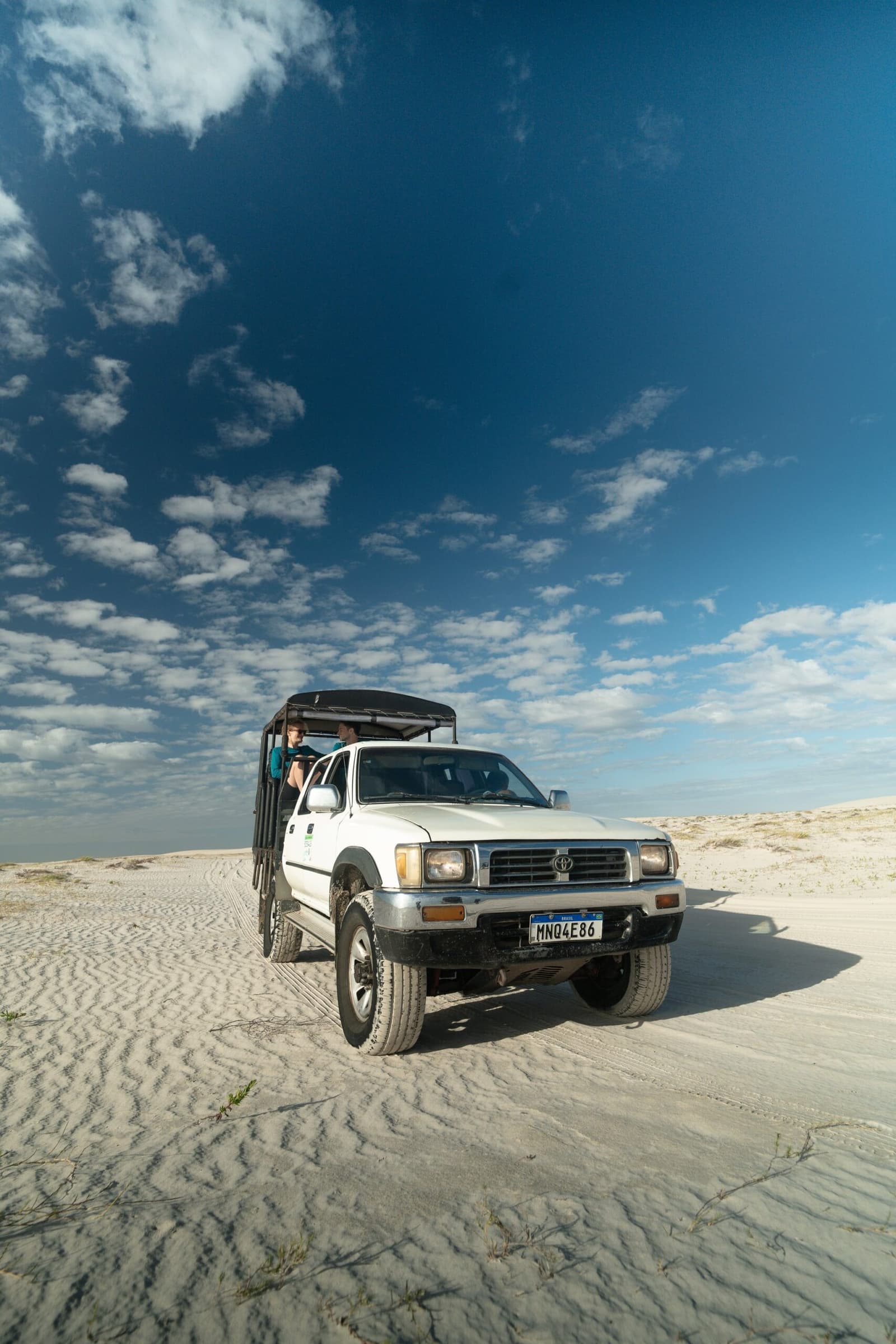 Your ride through the dunes: roof optional, views guaranteed. A white Toyota 4x4 pickup with an open cargo bed and passengers standing up crosses vast white sand dunes under a blue sky with scattered clouds, at the start of a Lençóis Maranhenses trekking journey