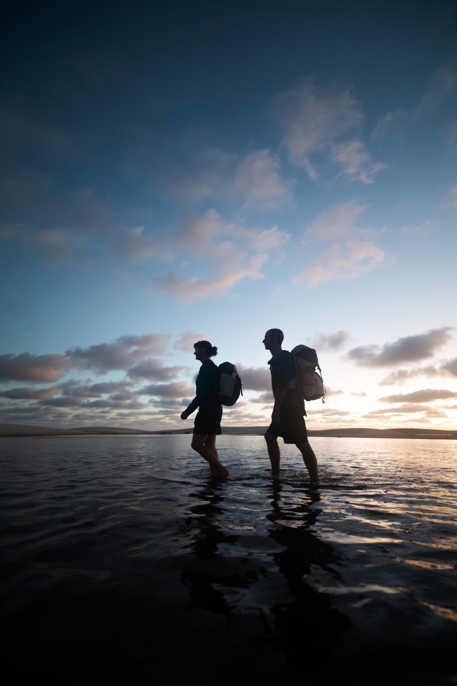 Wet feet, big skies, and nowhere else you'd rather be. Two hikers with backpacks silhouetted against a dusk sky, wading barefoot through shallow water at the end of a Lençóis Maranhenses trekking day
