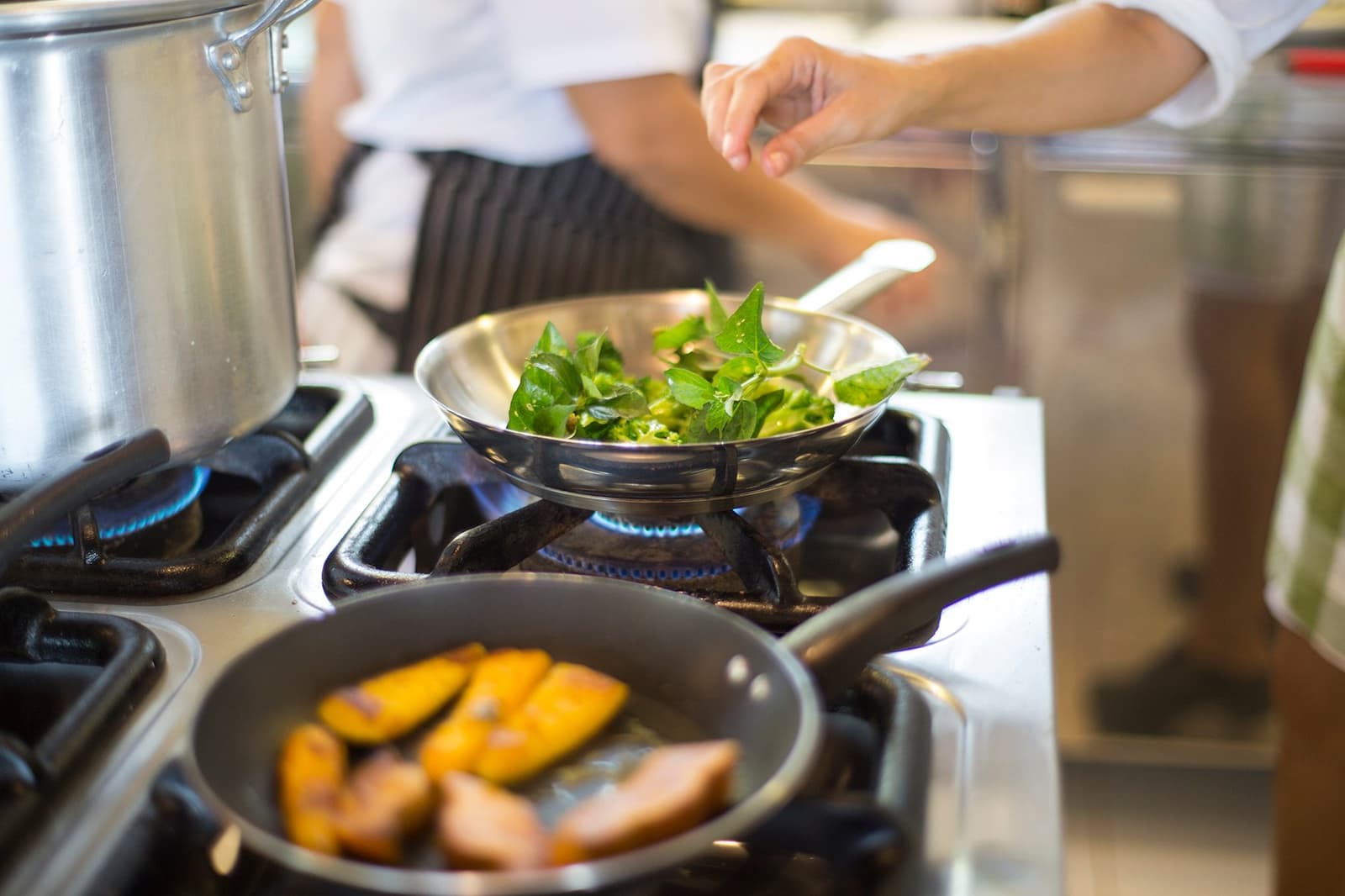 Lodge cooking that actually makes you want to skip the hammock. A chef in a striped apron sautéing fresh green leaves in a stainless steel pan over a gas flame at the Mirante do Gavião lodge kitchen, with golden-seared fruit slices cooking in a second pan in the foreground