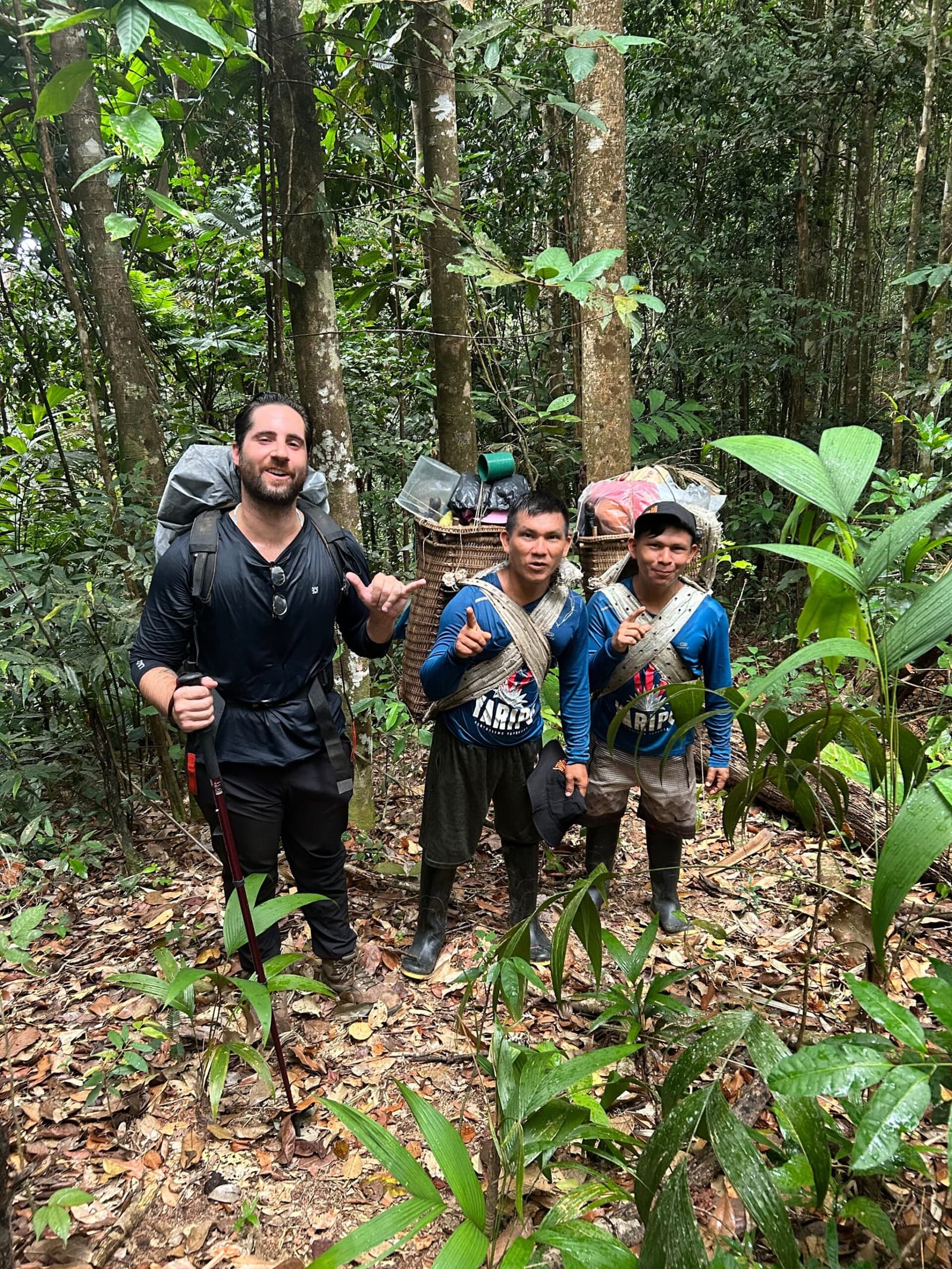 Three packs, one trail, and a good vibe that needs no translation. A traveler with a trekking pole and large backpack smiles alongside two Yanomami guides carrying heavily loaded traditional woven baskets on a Pico da Neblina Expedition trail through dense Amazon rainforest