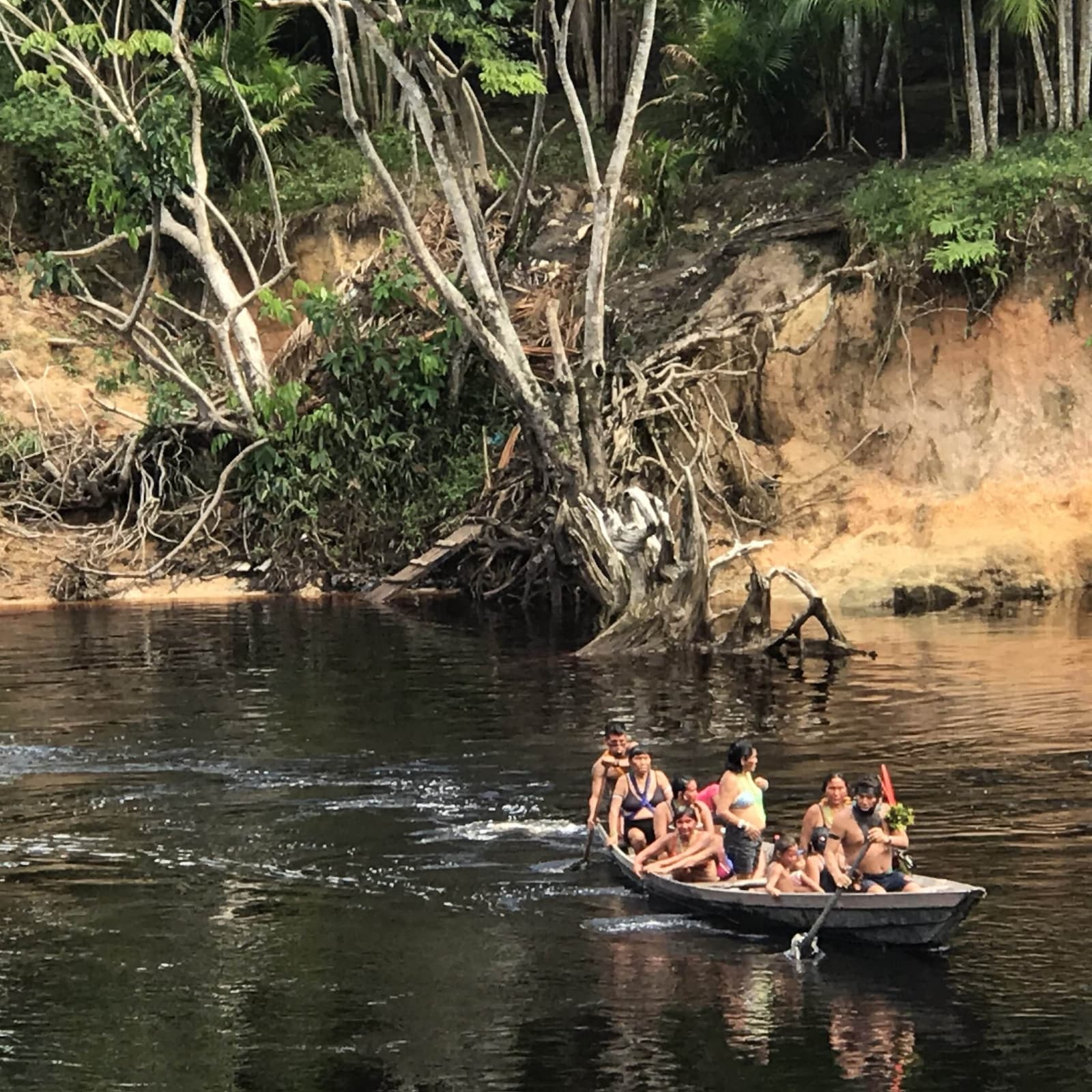 Local life on the river moves at its own pace, and always fits one more person. A crowded wooden motorized canoe carrying a local family across a dark-water Amazonian river, with an eroded clay riverbank and dense tropical forest rising behind them, during the Pico da Neblina Expedition.