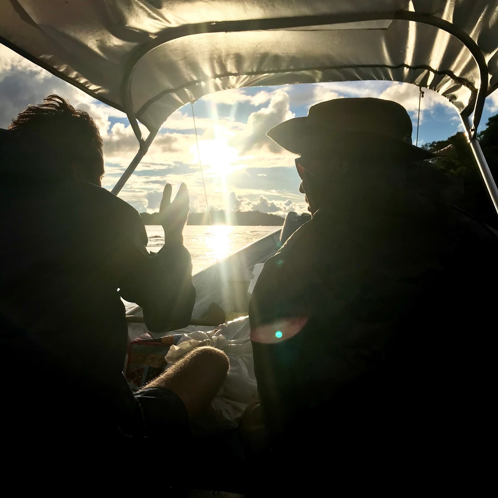 Peace signs and sun glare somewhere deep in the northwest Amazon. Two travelers silhouetted inside a covered motorized canoe on the Pico da Neblina Expedition, one flashing a peace sign as the sun bursts through clouds over a wide Amazonian river
