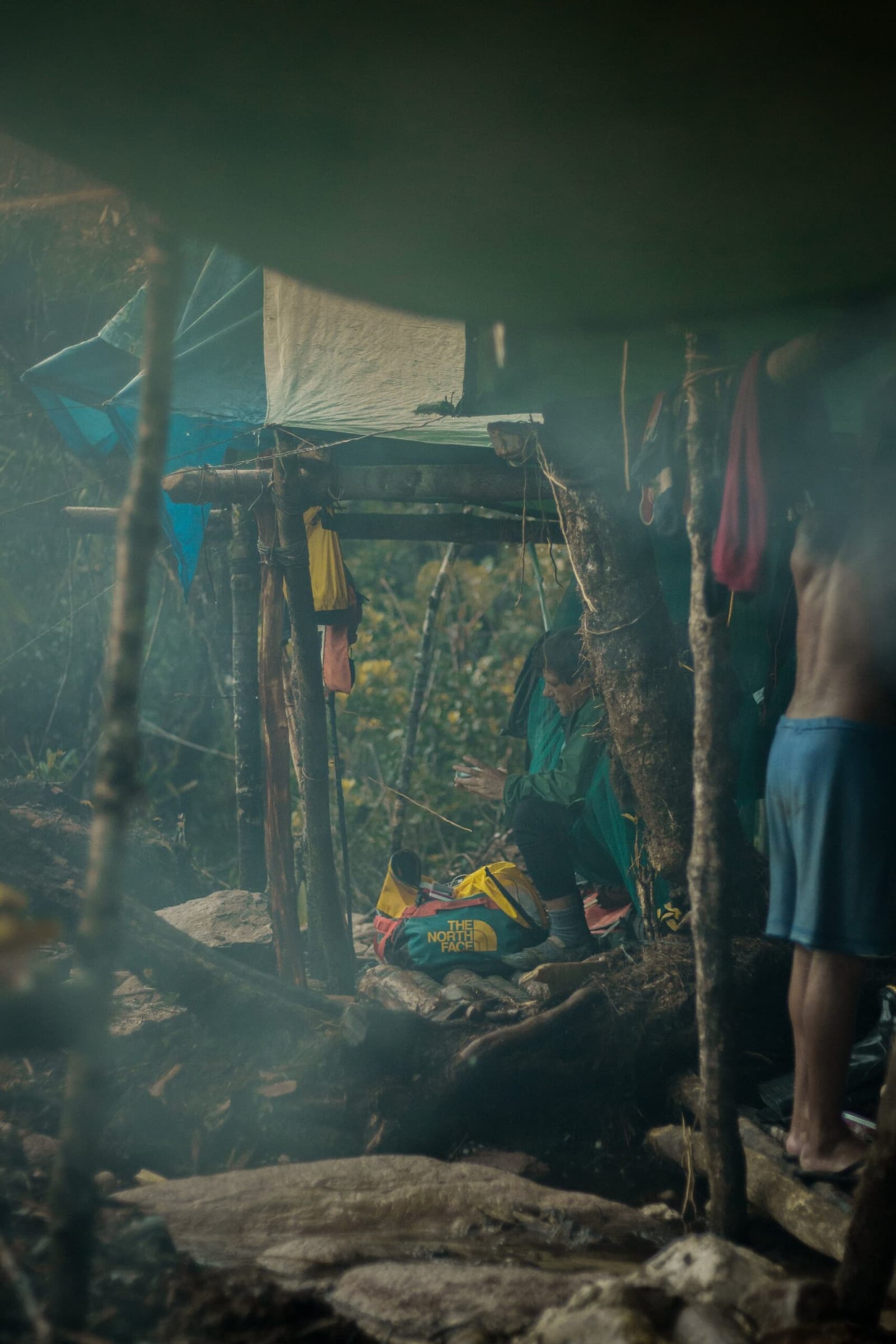 Smoke, altitude, and a bag that has seen better days. Camp life earns its reputation. A trekker crouches beside a yellow North Face dry bag inside a makeshift wooden shelter during the Pico da Neblina Expedition, smoke drifting through the hazy air while colourful gear hangs from bamboo poles