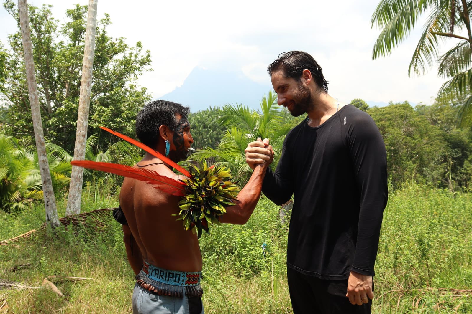 A handshake that carries more weight than most. A Yanomami man wearing traditional face paint, red feathers and a leaf adornment clasps hands warmly with a traveler during the Pico da Neblina Expedition, with tropical forest and a mountain peak visible in the background.