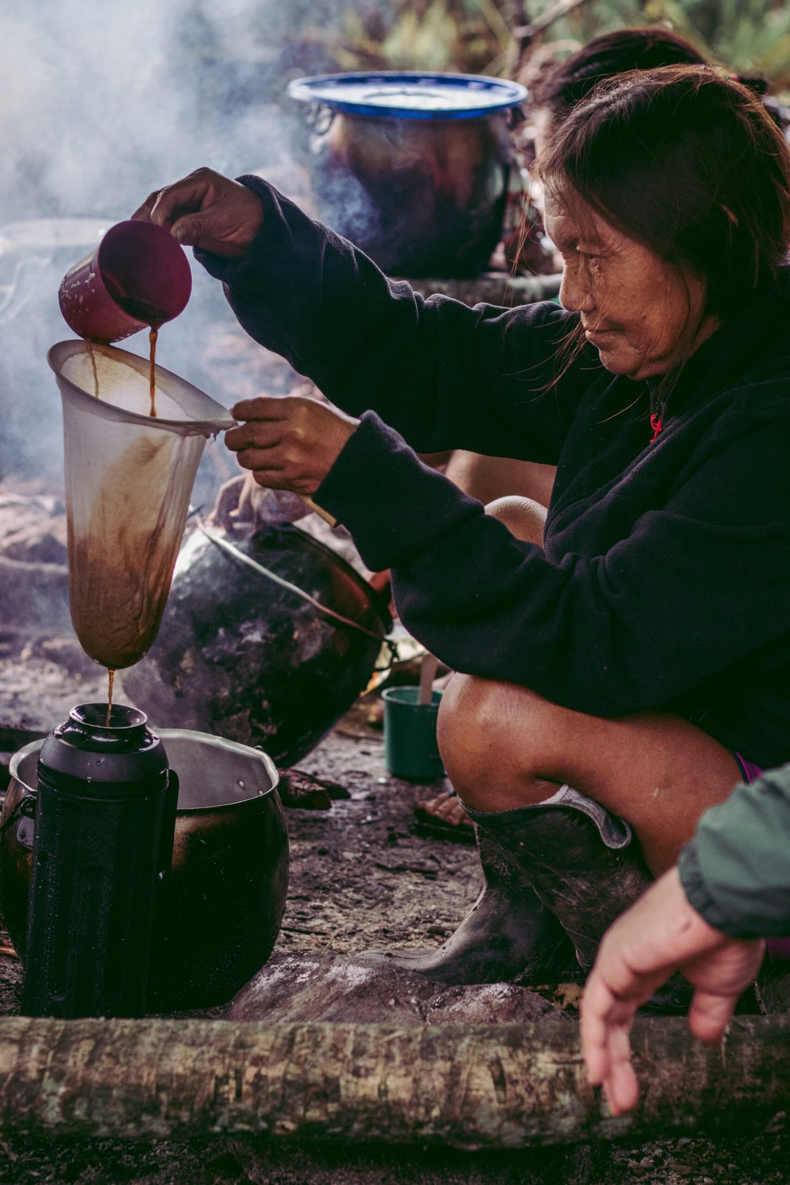 Coffee filtered by hand, fire at her back, smoke in the air. Some routines just work. A Yanomami woman crouches by a smoky open fire, carefully pouring dark liquid through a cloth filter into a blackened kettle during the Pico da Neblina Expedition