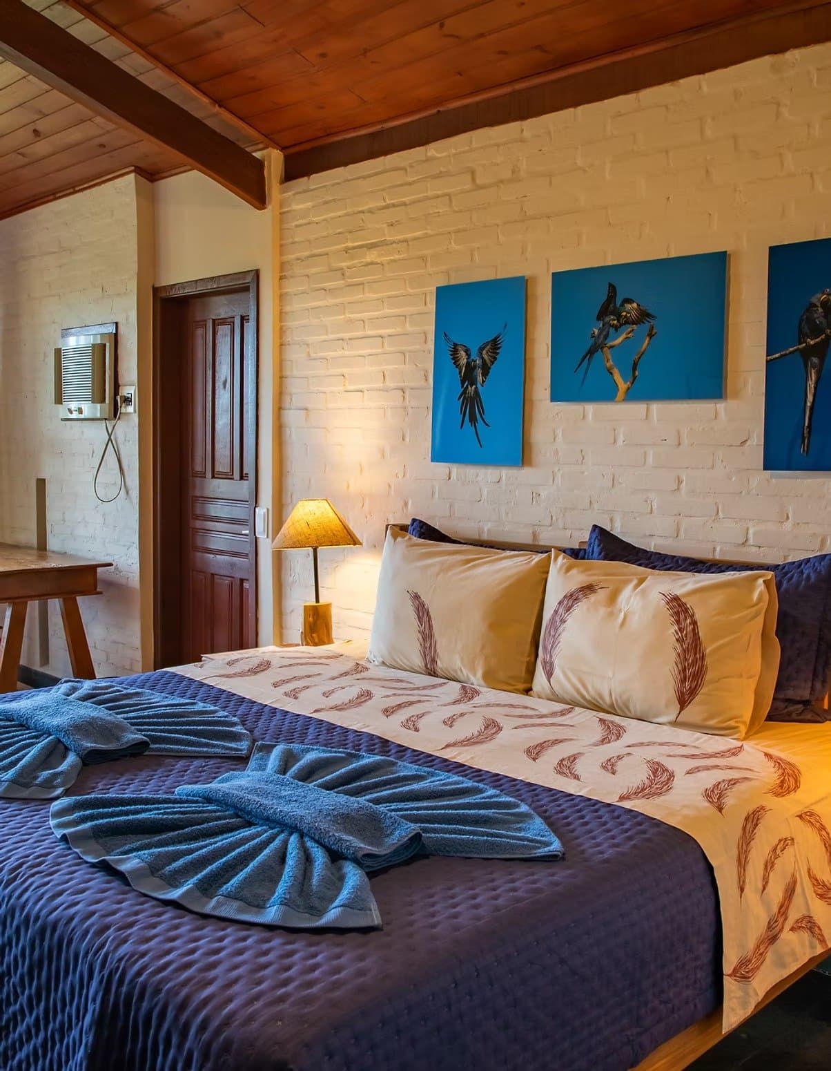 Macaws on the wall, feathers on the pillow. The lodge knows its audience. A southern pantanal safari lodge bedroom with a double bed dressed in feather-print linen, blue quilted coverlet, and towels folded into a fan shape, beneath three vivid blue canvases depicting macaws, warm lamplight, and a white-painted brick wall with a wood-beam ceiling