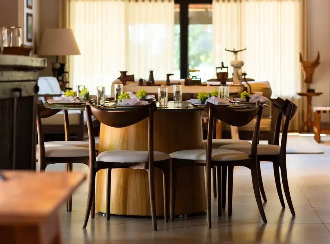 Dinner after a full day in the wetlands hits differently at a table set like this. Elegantly set round dining table with folded linen napkins, glassware, and small green floral arrangements in a warm southern pantanal safari lodge interior with vintage wooden furnishings and sheer curtains