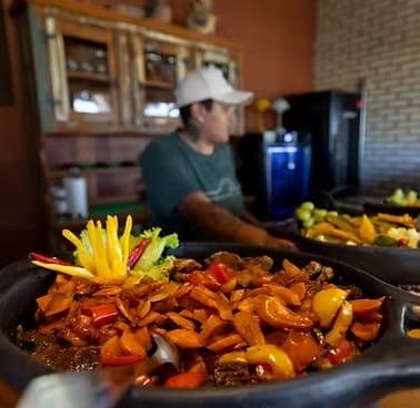 Pantaneiro cooking doesn't hurry, and neither should you. A cast-iron pan heaped with slow-cooked meat, bell peppers, and onions garnished with julienned yellow pepper, served at a southern pantanal safari lodge while a cook works in the background