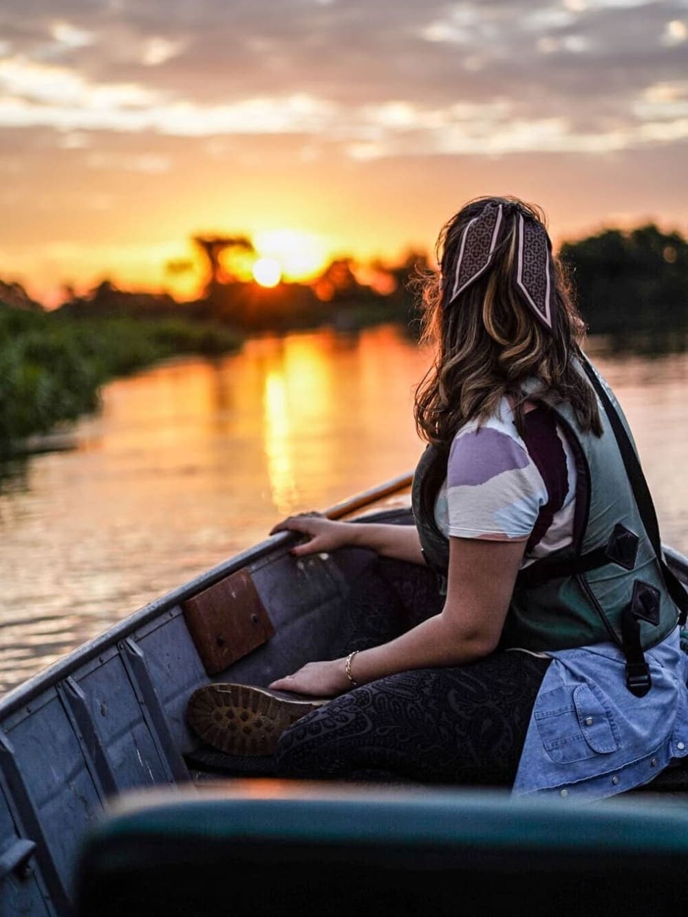 Golden hour hits different when the only engine noise is your own paddle. A woman wearing a life jacket sits in a small wooden canoe, paddle in hand, watching the sun set over a calm river channel flanked by low vegetation on a southern pantanal safari