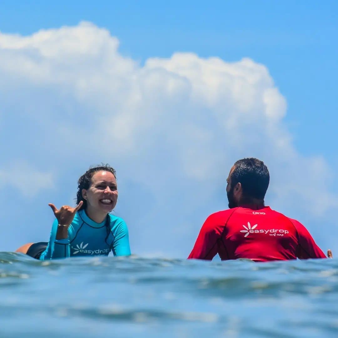 The shaka says it all, some lessons click faster than expected. A smiling student in a teal rash guard flashes a shaka sign to her instructor in red at the surf school in Itacaré, both sitting on their boards beyond the break under a bright blue sky.