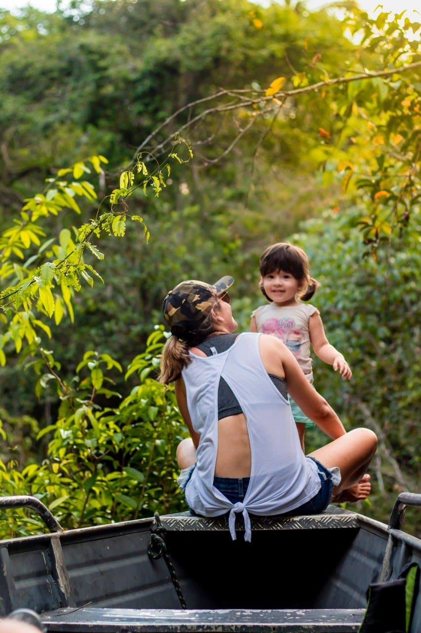 Kids have a way of making every jungle transfer feel like the best part of the trip. A woman in a camo cap and a smiling toddler sit together on the back of a vehicle surrounded by lush Amazon rainforest vegetation during an Amazon boutique lodge excursion