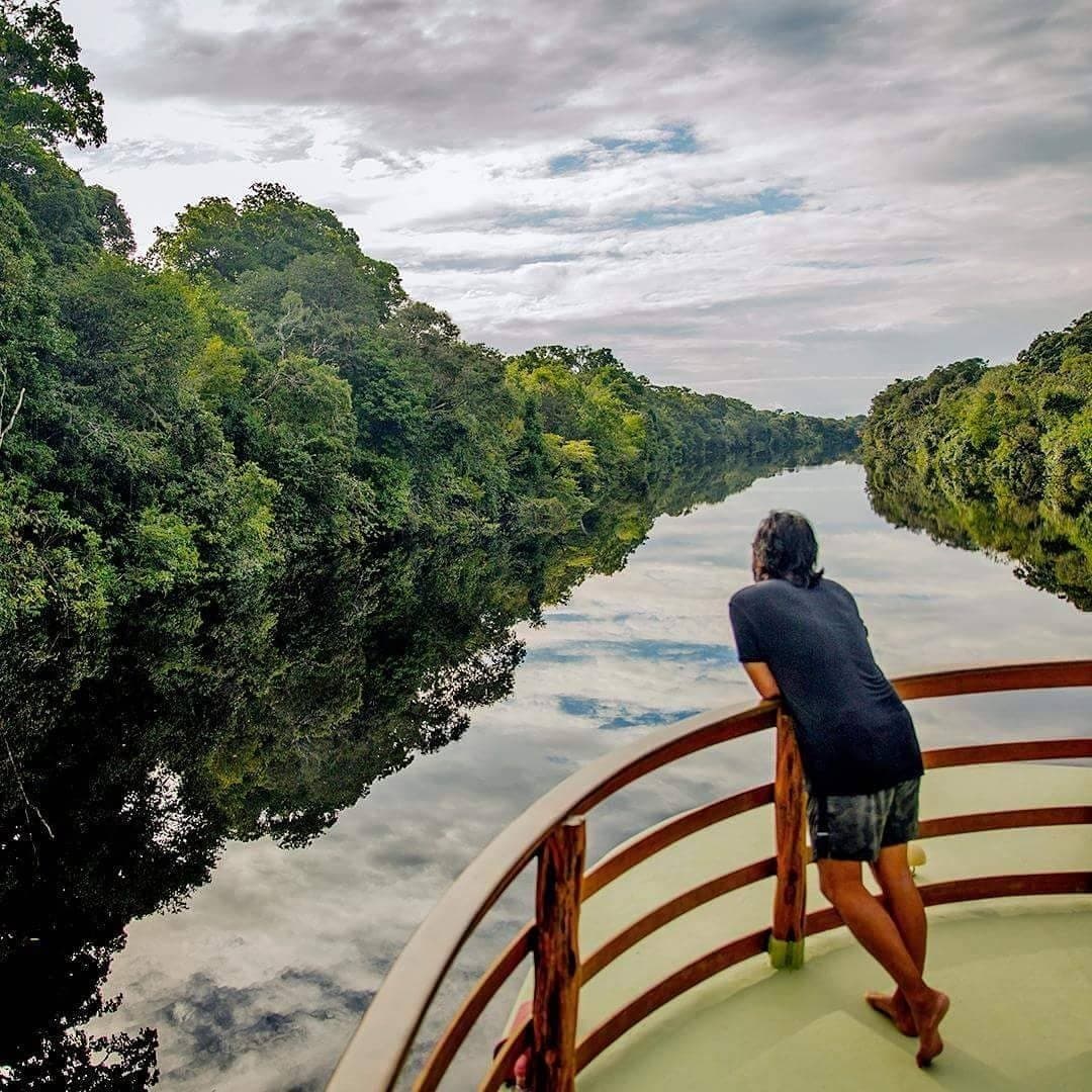 The river does all the talking, you just have to lean in and listen. A traveller leans on the curved bow railing of a river cruise boat, gazing along a glassy jungle-lined waterway on an Amazon jungle cruise Brazil — dense rainforest canopy reflected perfectly in the still dark water beneath a cloudy sky