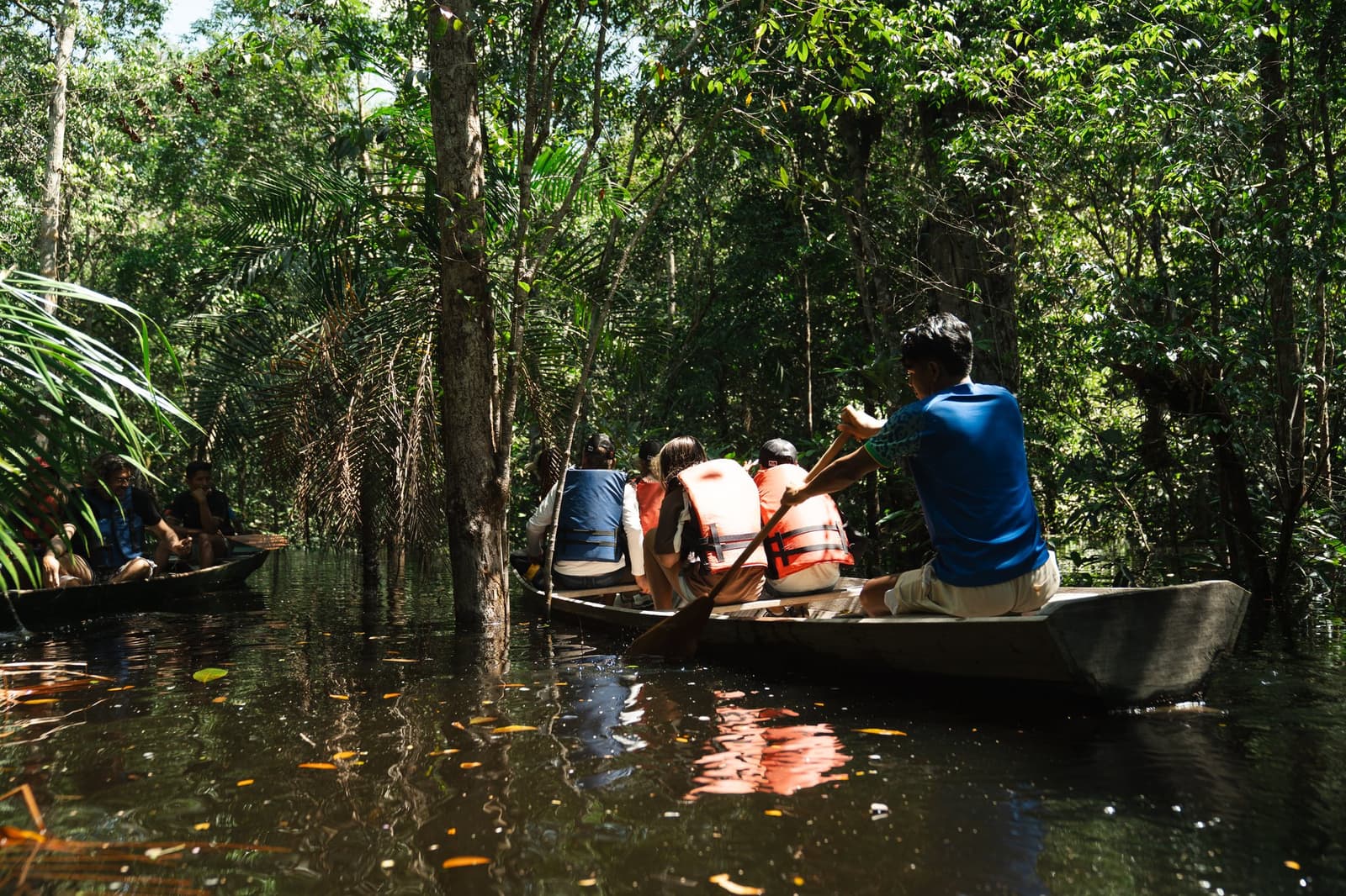The forest opens up differently when you're sitting two feet above the water. Guests in life jackets paddling wooden dugout canoes through a flooded igapó forest on an amazon turtle lodge rainforest excursion