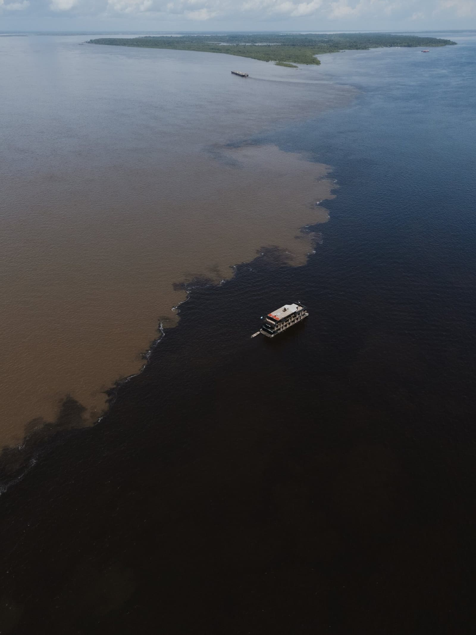 Two rivers, two colours, one line, and your boat right in the middle of it. Aerial view of a river boat crossing the Meeting of Waters on an amazon rainforest vacation brazil, where dark black-water and sandy brown-water rivers meet in a vivid swirling boundary, with a forested island in the distance