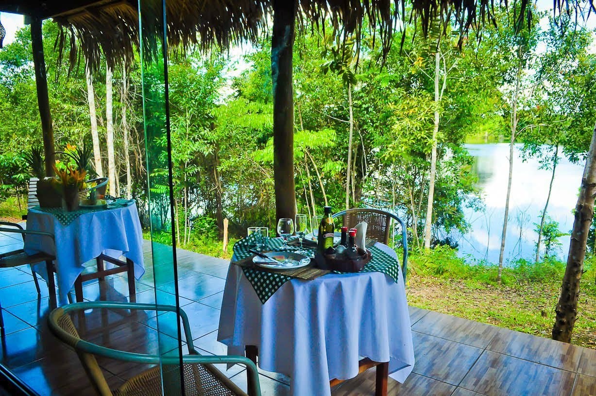 Dinner with the river as your only neighbour. Open-air thatched dining terrace at Amazon Turtle Lodge with white-clothed tables set for dinner, wine bottles, and a tranquil river framed by dense rainforest trees