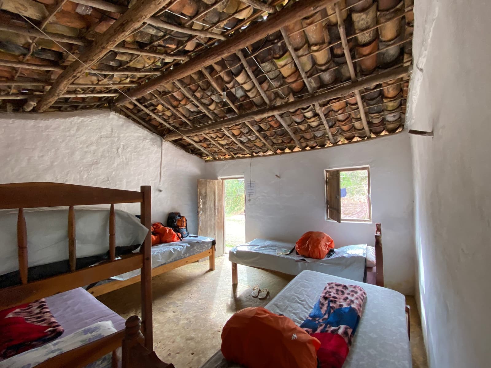 Four walls, honest beds, and the valley waiting outside the door. Simple shared bedroom in a Vale do Pati family homestay, with wooden bunk beds and single beds topped with trekkers' orange dry bags, exposed clay roof tiles on wooden beams, whitewashed walls, and a small window opening onto greenery outside — typical Chapada Diamantina trek accommodation