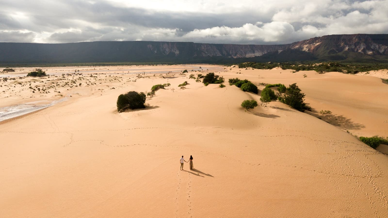 Just two people, a whole lot of sand, and footprints going exactly where they wanted. Photo: FG Trade Aerial view of a couple holding hands on the vast golden Jalapão sand dunes, with green shrubs, a river edge, and a dramatic mesa escarpment under a stormy sky — a quiet moment on a Jalapão adventure.