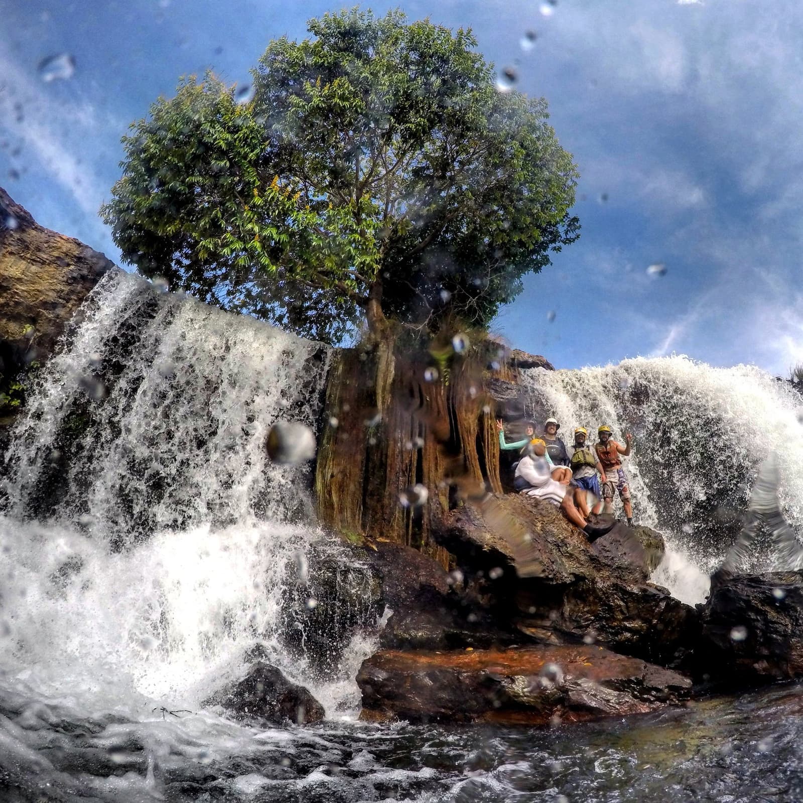Soaked, helmeted, and grinning from ear to ear, this is what rafting in Jalapão looks like up close. A group of travelers in helmets and life vests pose on rocks between two cascading waterfalls during a Jalapão adventure, with water droplets caught mid-air by the camera lens and a lone tree rising above the falls against a blue sky