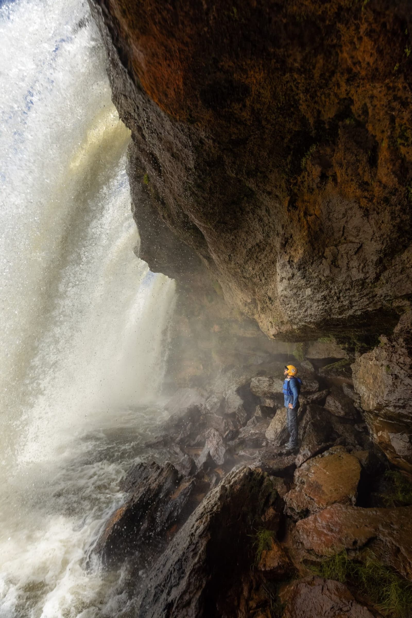 Standing inside the roar, Cachoeira da Velha puts you right in it. Photo: Alexandre Socci A hiker in a blue jacket and orange helmet stands on wet rocks beneath a massive waterfall overhang, looking up at the thundering cascade on a Jalapão trekking route
