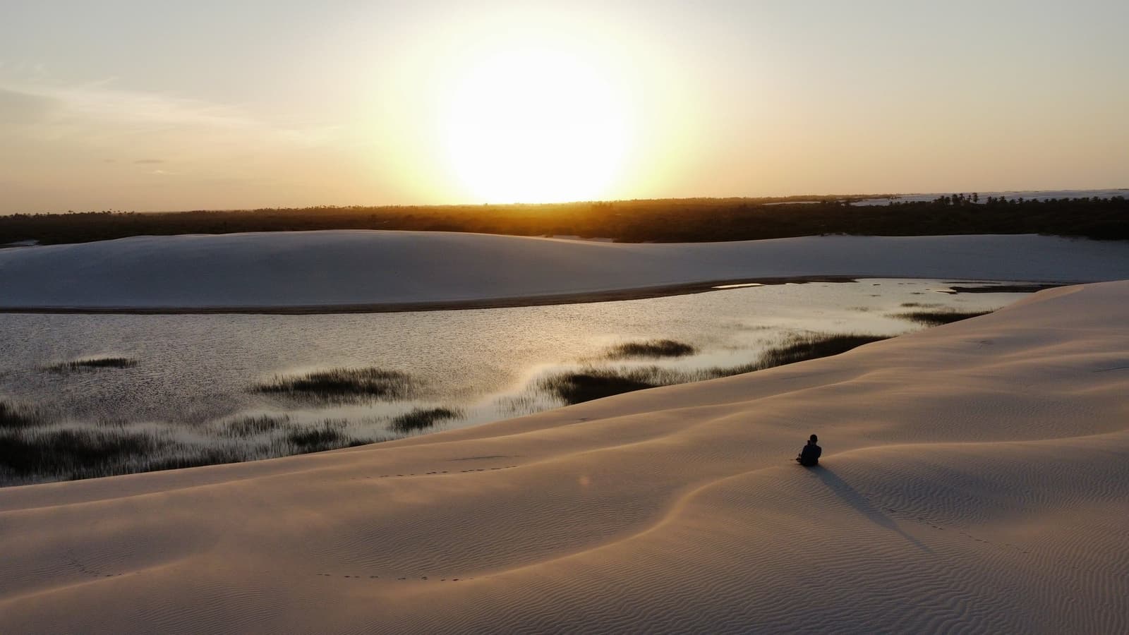 One person, one dune, and the whole evening to themselves. A lone figure sits on rippled white sand dunes overlooking a shallow lagoon fringed with grass tufts at golden sunset, a defining scene of the Lençóis Maranhenses full itinerary